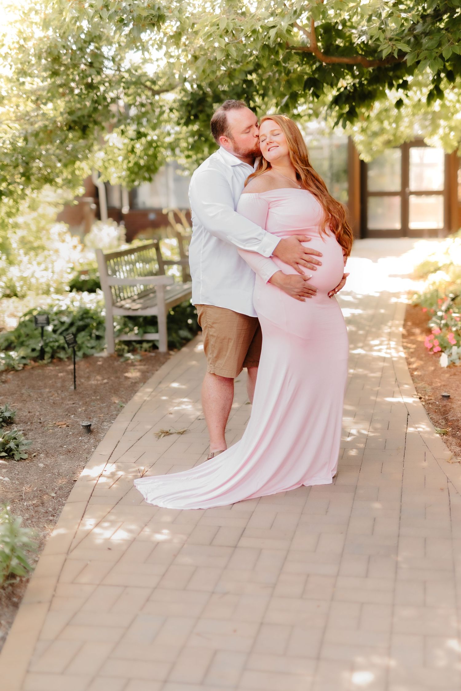 A couple shares a tender moment together on a garden pathway as sunlight filters through the trees overhead.
