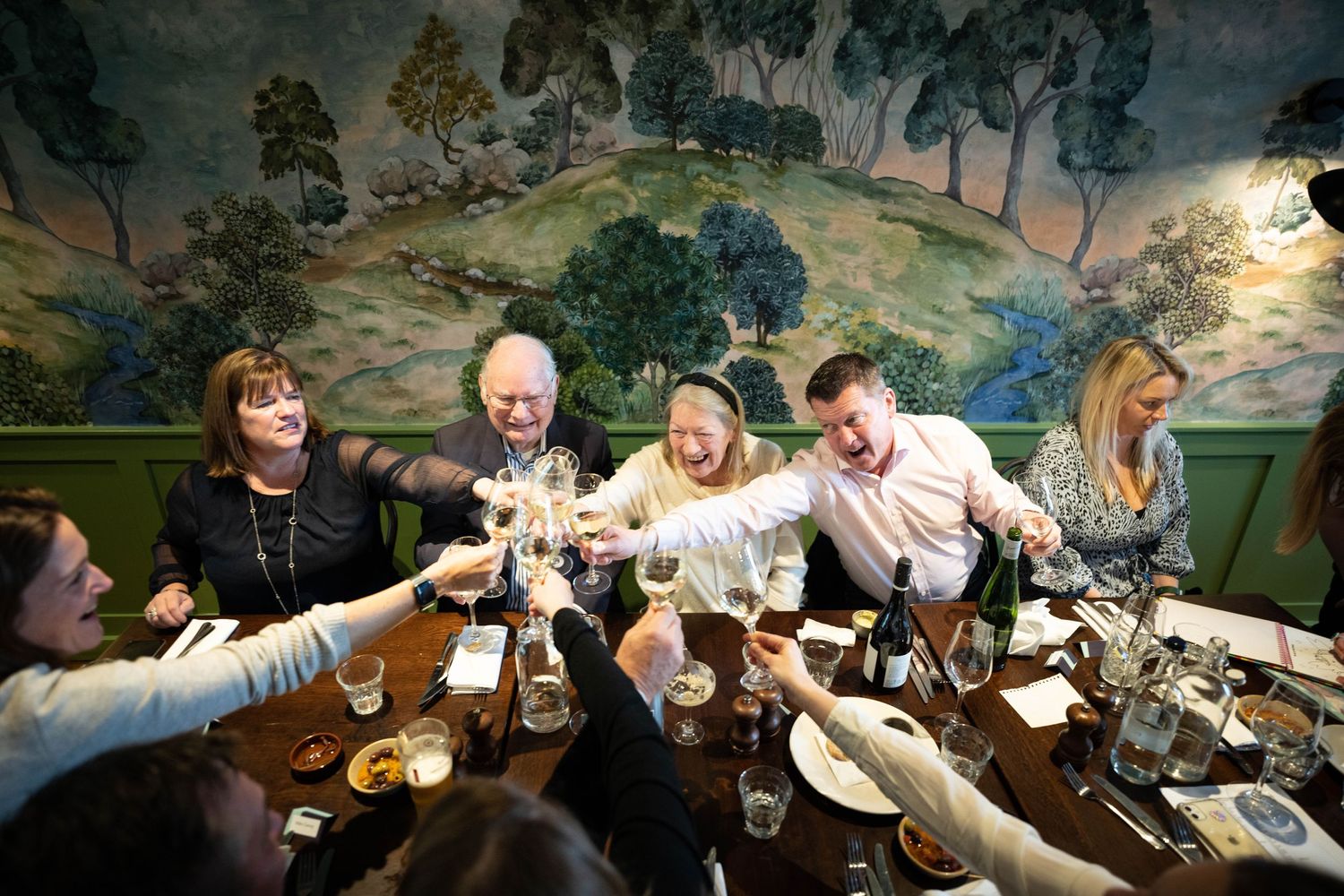 Dinner party guests toast around table with decorative pastoral mural backdrop.