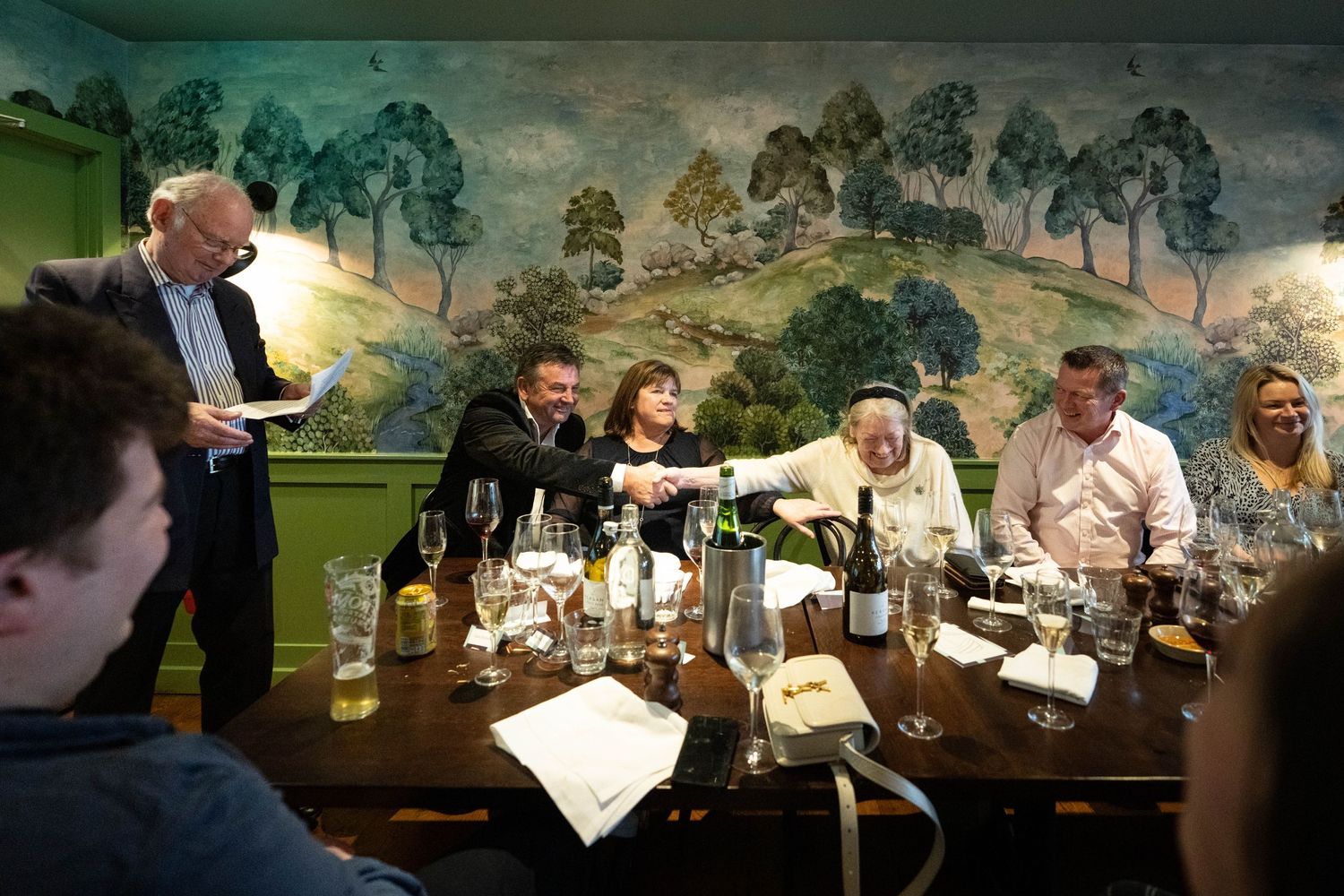 A group dining together at a restaurant with a scenic pastoral mural on the wall in the background.