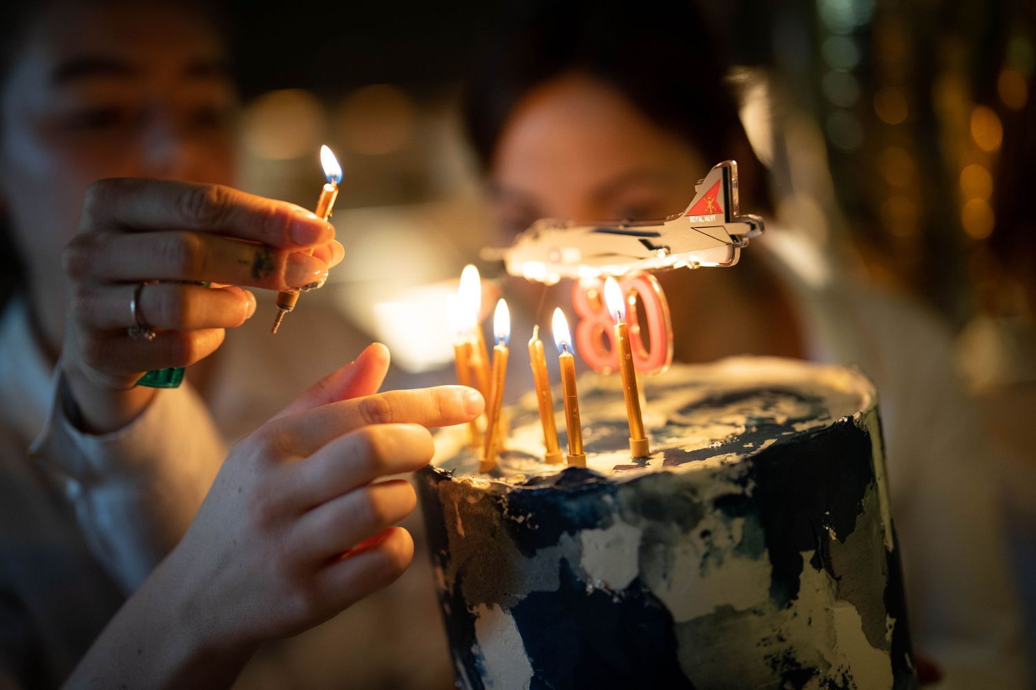 Close-up of hands lighting candles on a dark-colored birthday cake.