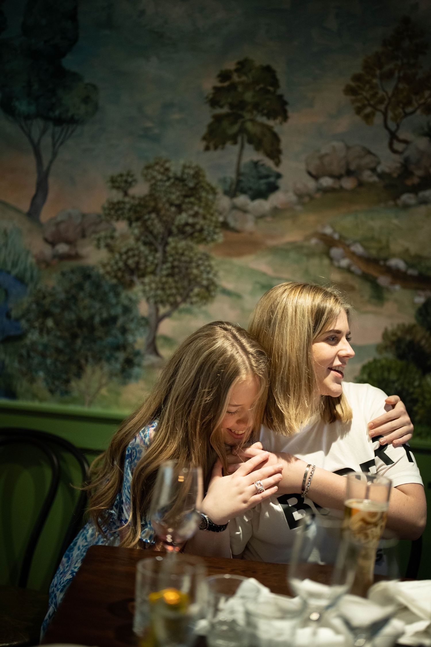 Two guests sharing a joyful moment at a dinner party with a painted landscape backdrop.