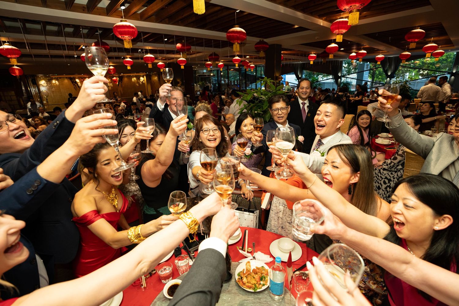 Large group toasting with raised glasses at a festive banquet under red lanterns.