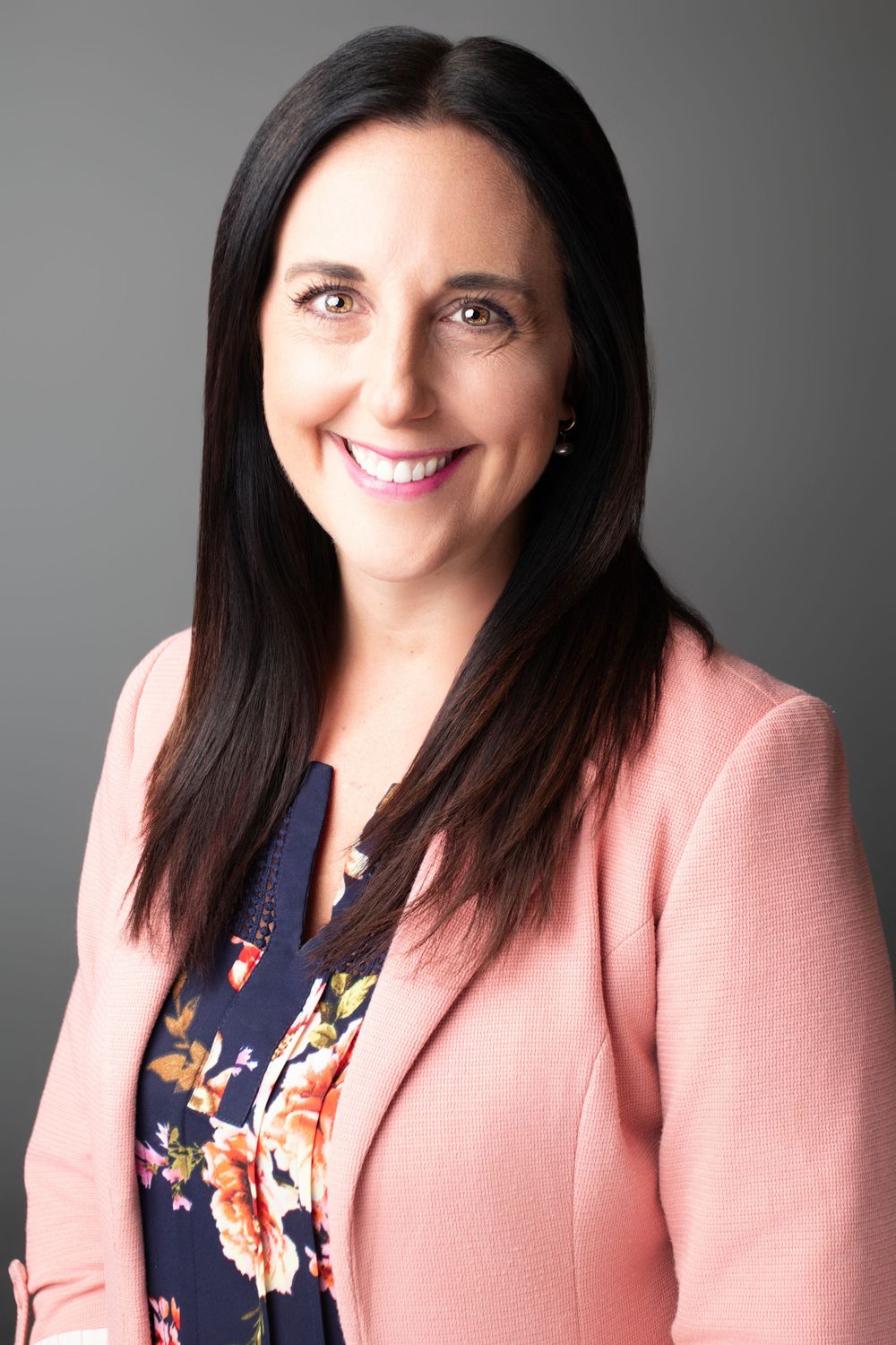 Professional headshot of woman in pink blazer and floral top against gray background.