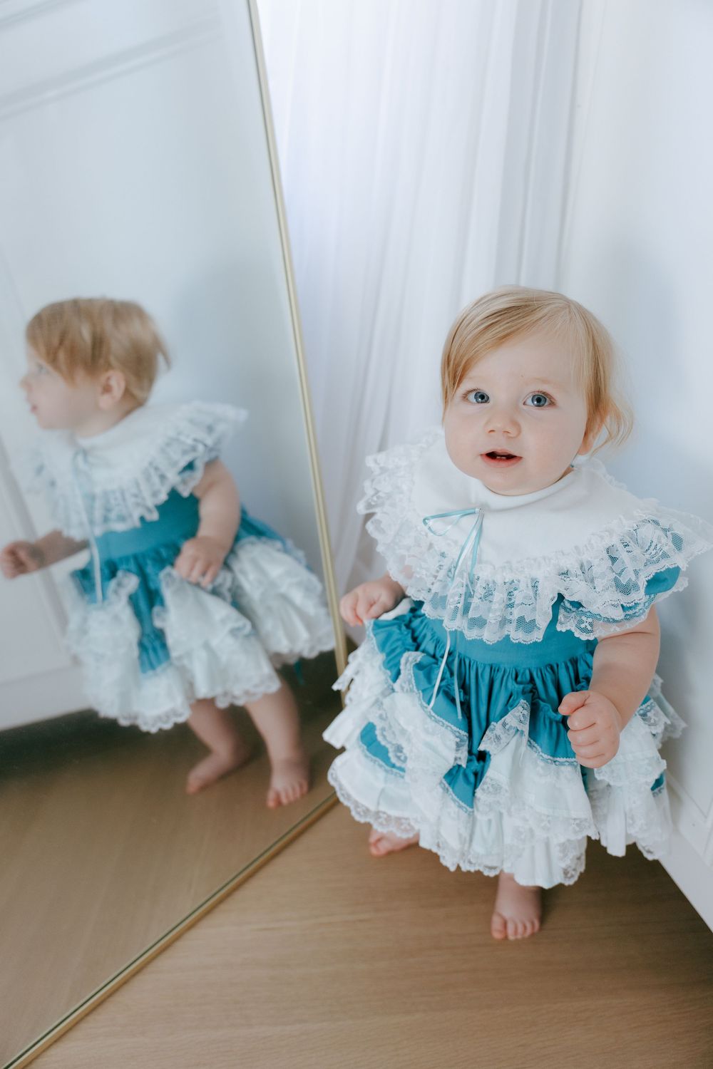 Baby in turquoise and white lace dress stands near mirror reflection on hardwood floor.
