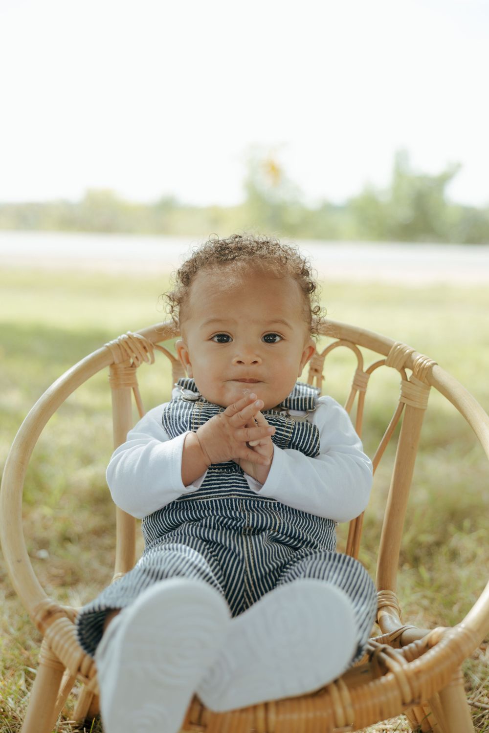 Small child wearing a bowtie and overalls sits in a woven rattan chair outdoors on a sunny day.