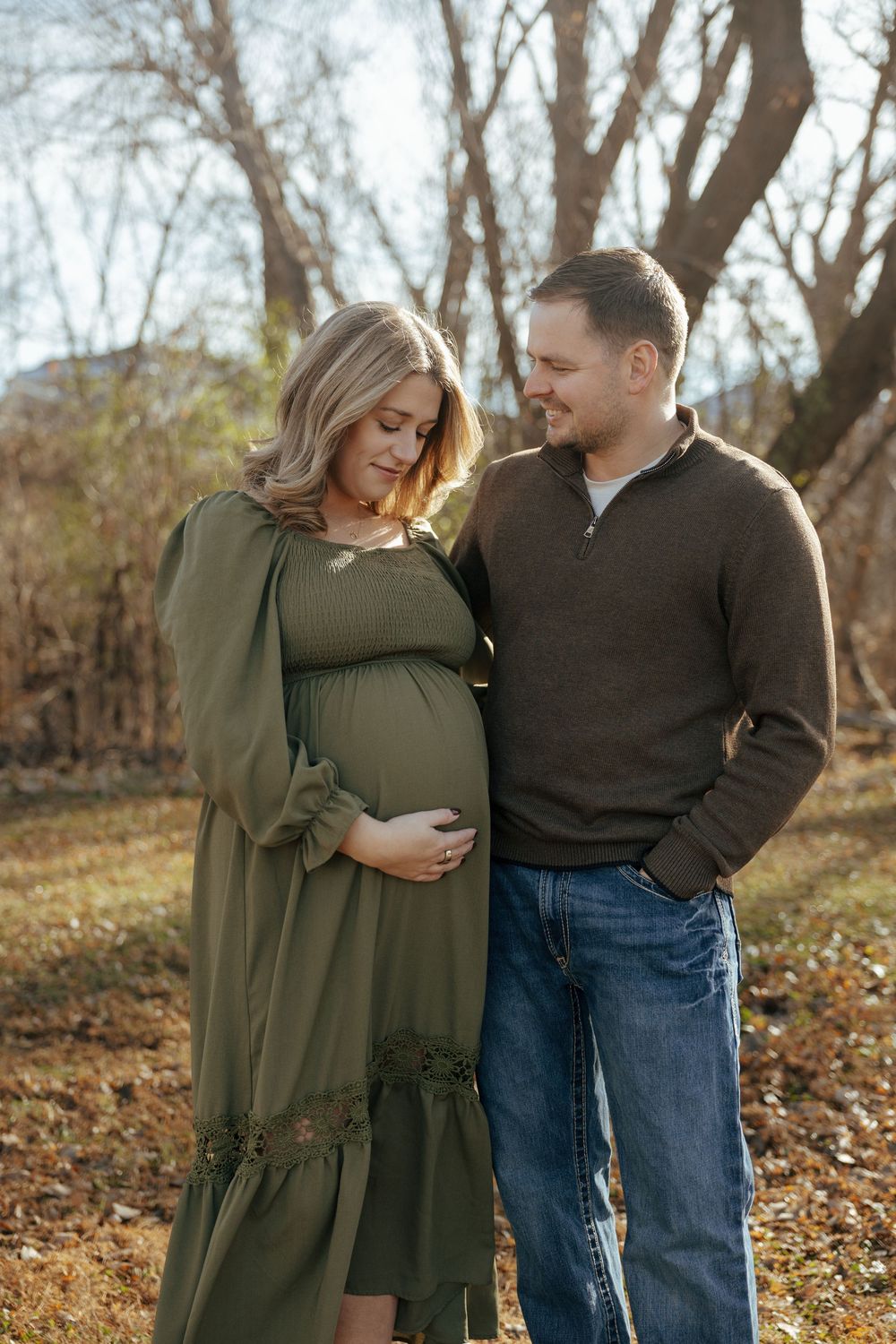 A couple in casual attire embraces while standing among fallen autumn leaves in a wooded area.