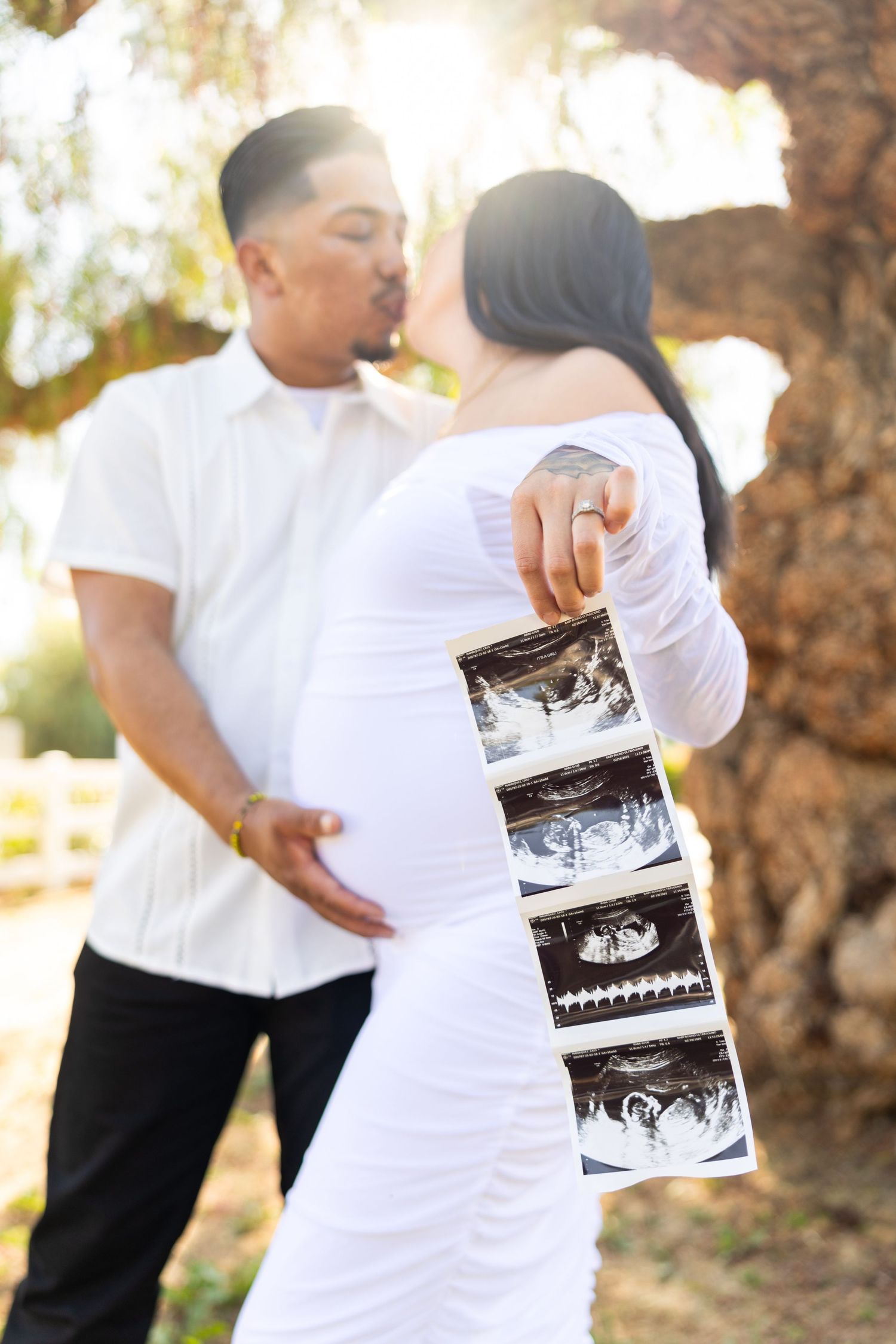 A couple in white outfits stands outdoors holding ultrasound photos during a pregnancy announcement photoshoot.