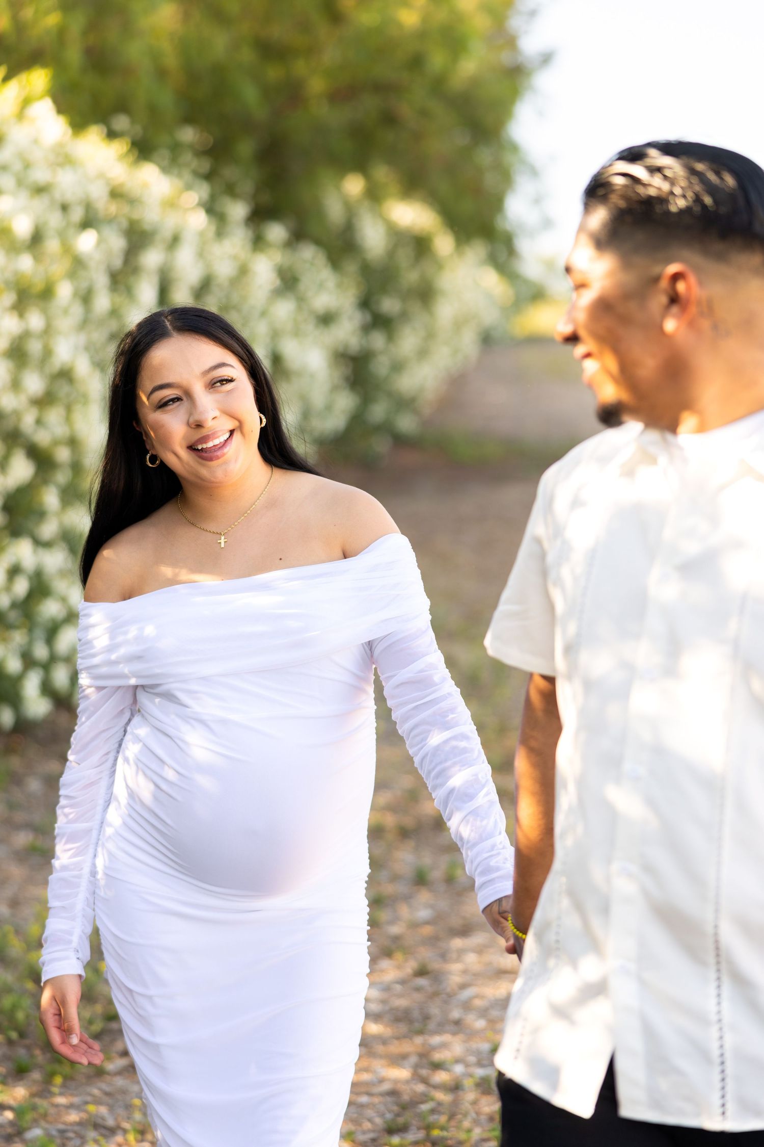 A couple walks along a nature path with white flowers blooming, the woman wearing a white off-shoulder maternity dress.