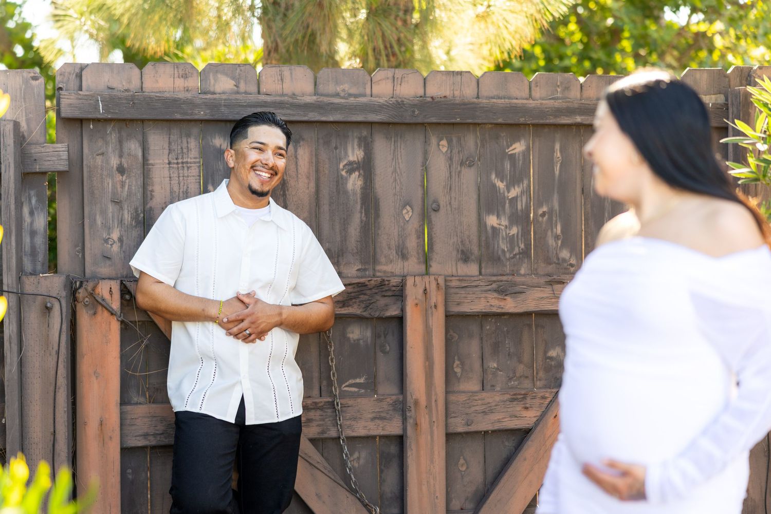 A couple in white clothing smile at each other during a maternity photoshoot by a rustic wooden fence in bright sunlight.