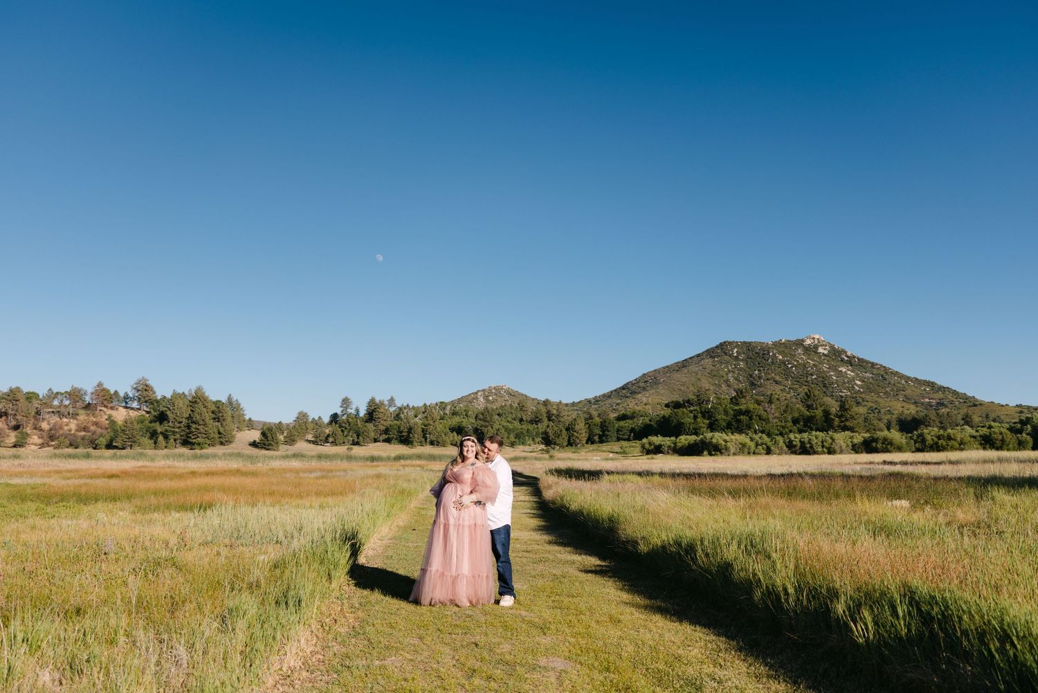 Two people walk hand in hand along a grassy path with a mountain and blue sky in the background.