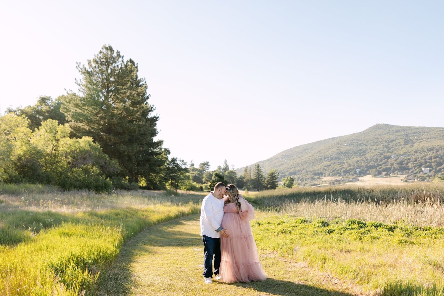 A couple walks hand in hand down a winding dirt path through golden grass with mountains in the background at sunset.