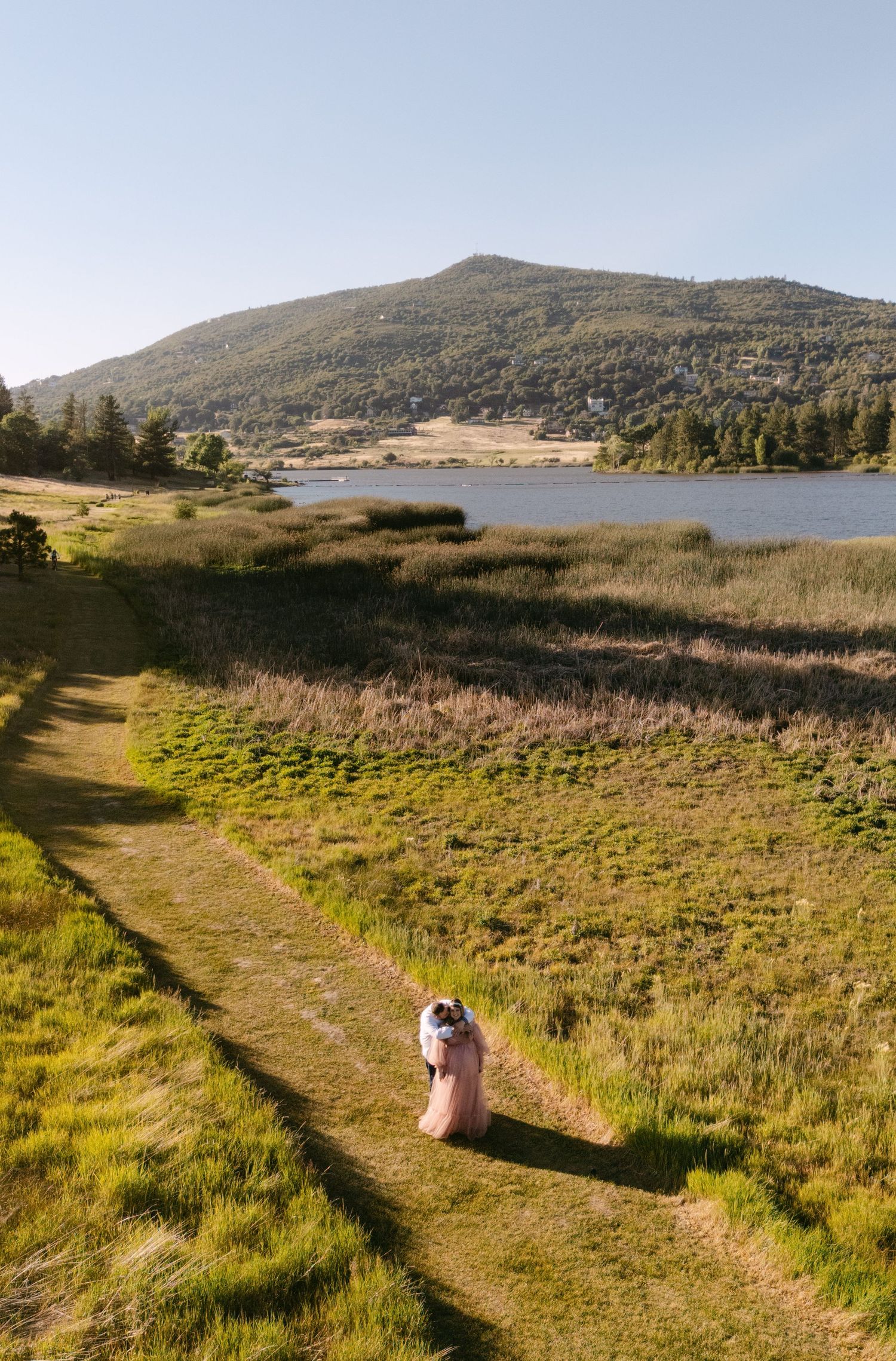A dirt hiking trail winds along a serene lake surrounded by hills and grassland on a sunny day.
