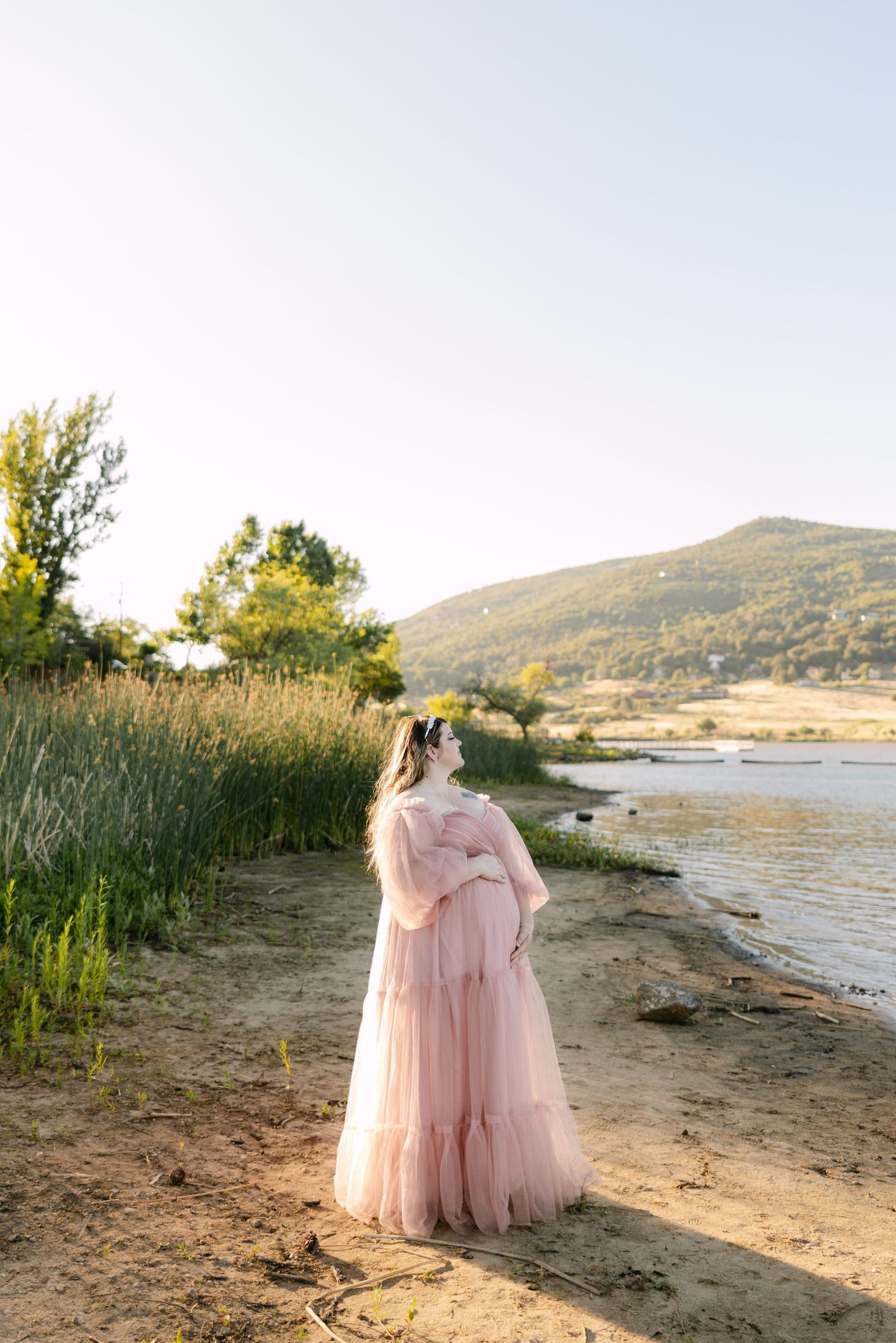 A pink tulle maternity dress catches golden sunlight on a serene lakeshore with mountains in the background.