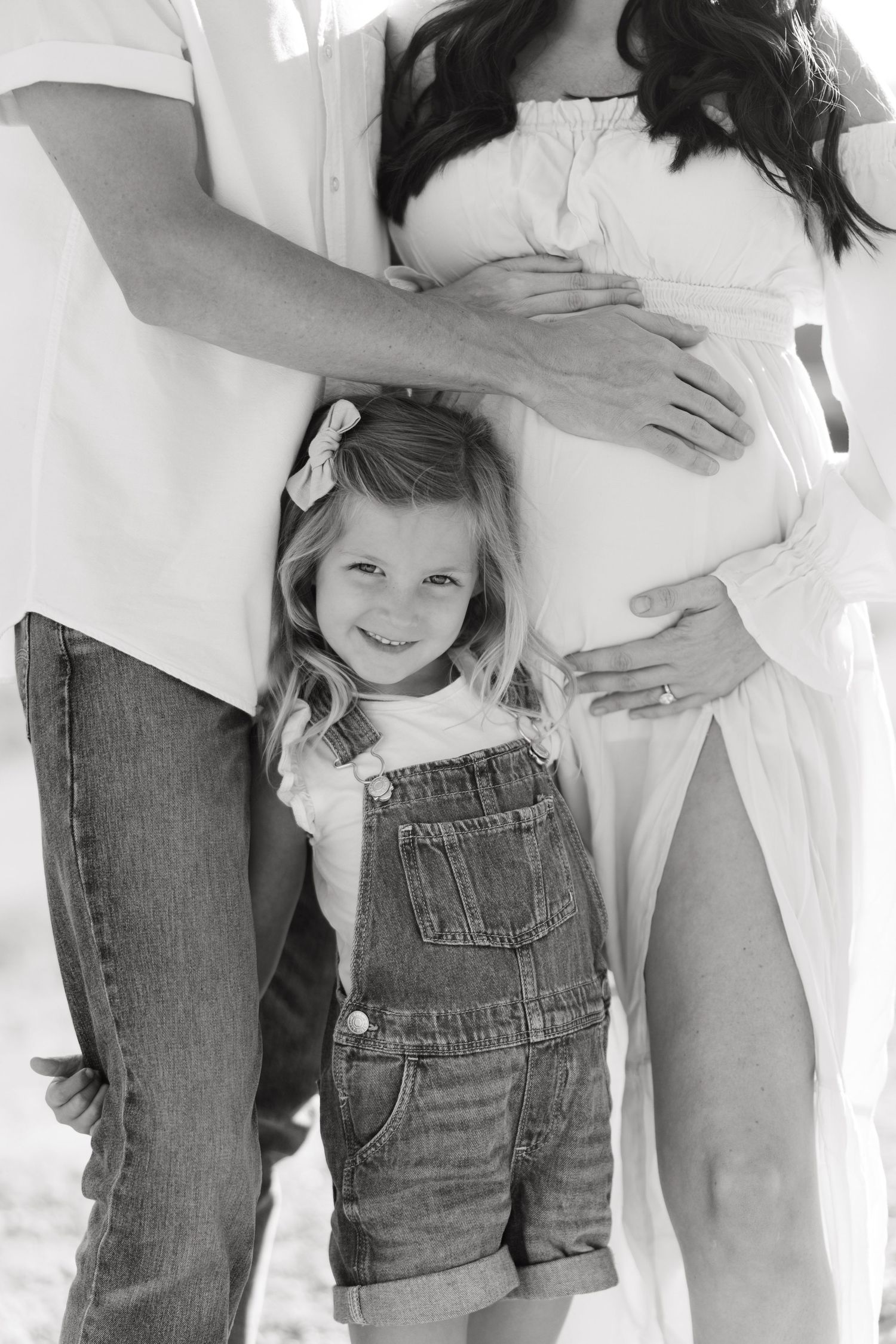 A black and white photo of a child in denim overalls standing between two people during a maternity photoshoot.