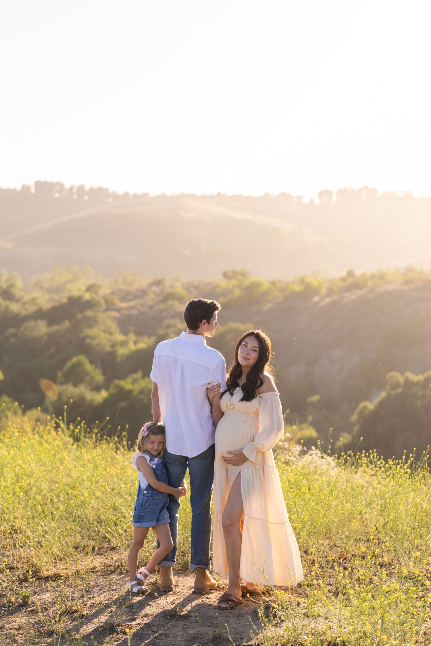 Family standing together on a hilltop at sunset in a golden field with rolling hills in the background.