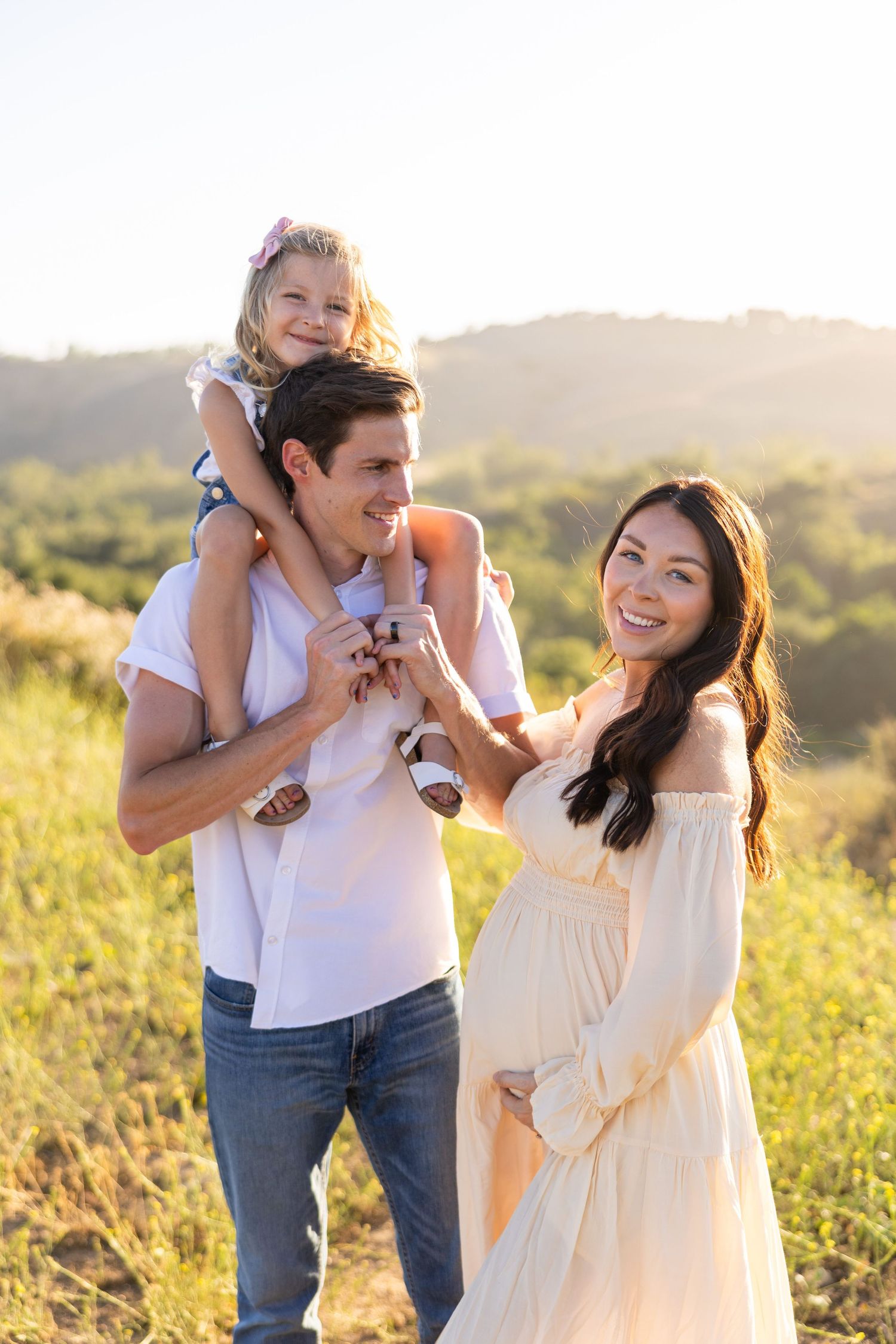 A couple in white outfits share joyful moments against a sunlit hilltop setting with golden grass in the background.