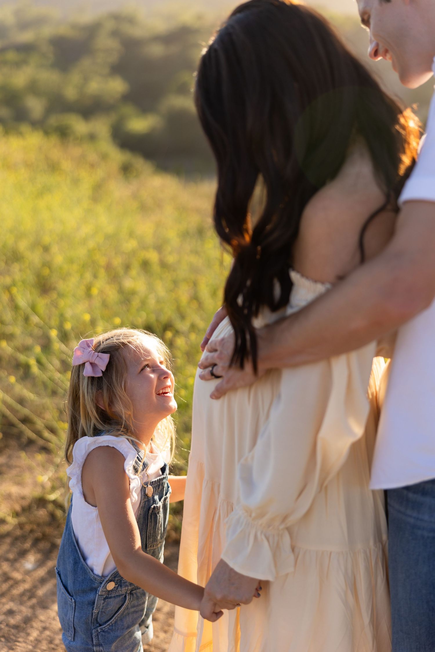 Young child in overalls holds hands with two adults during golden hour sunset in a grassy field.
