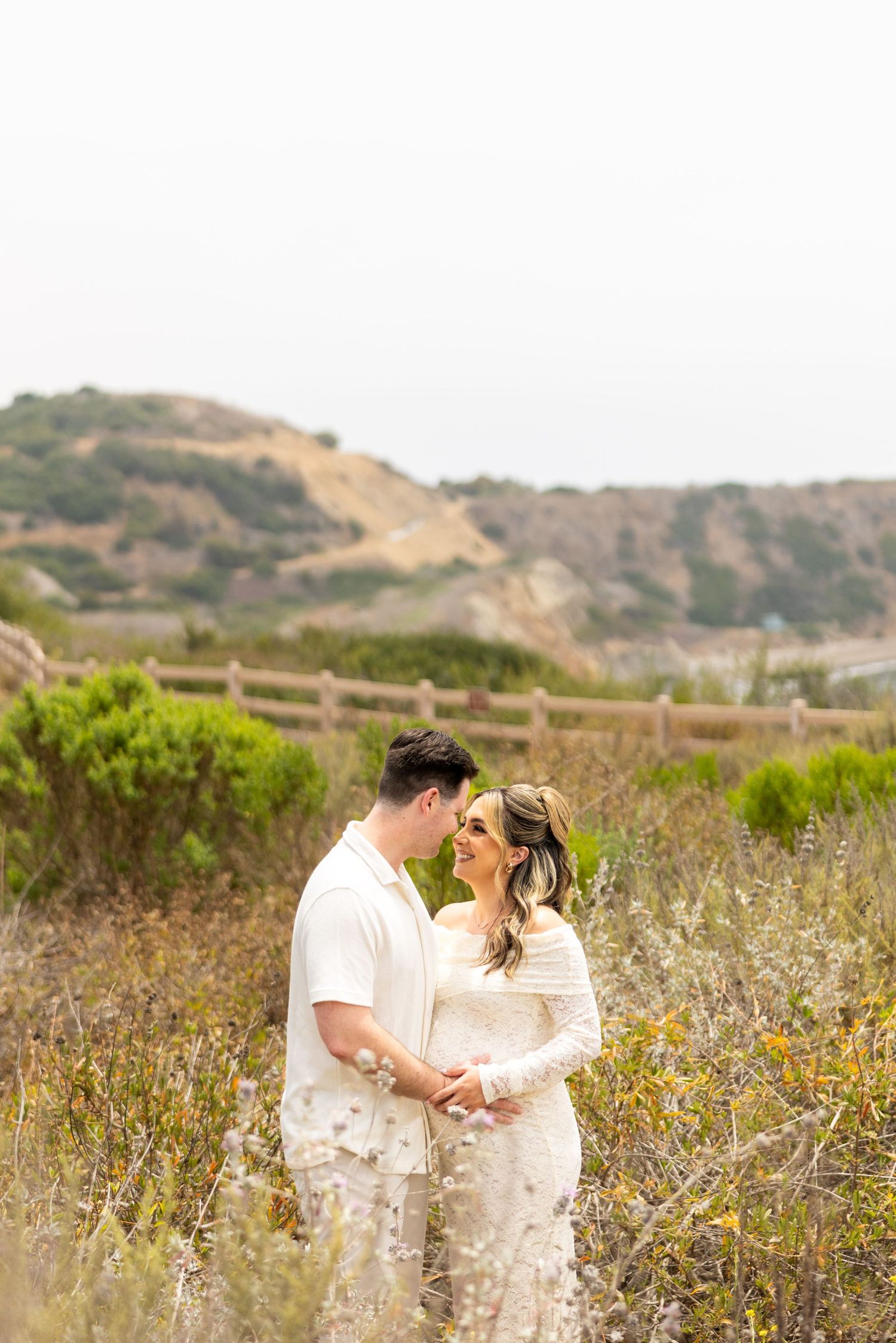 A couple shares a tender moment among wildflowers on a hillside with wooden fencing and coastal cliffs in the background.