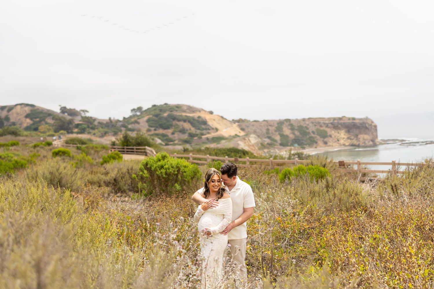 Two people embrace on a coastal cliff with wildflowers and ocean views in the background.