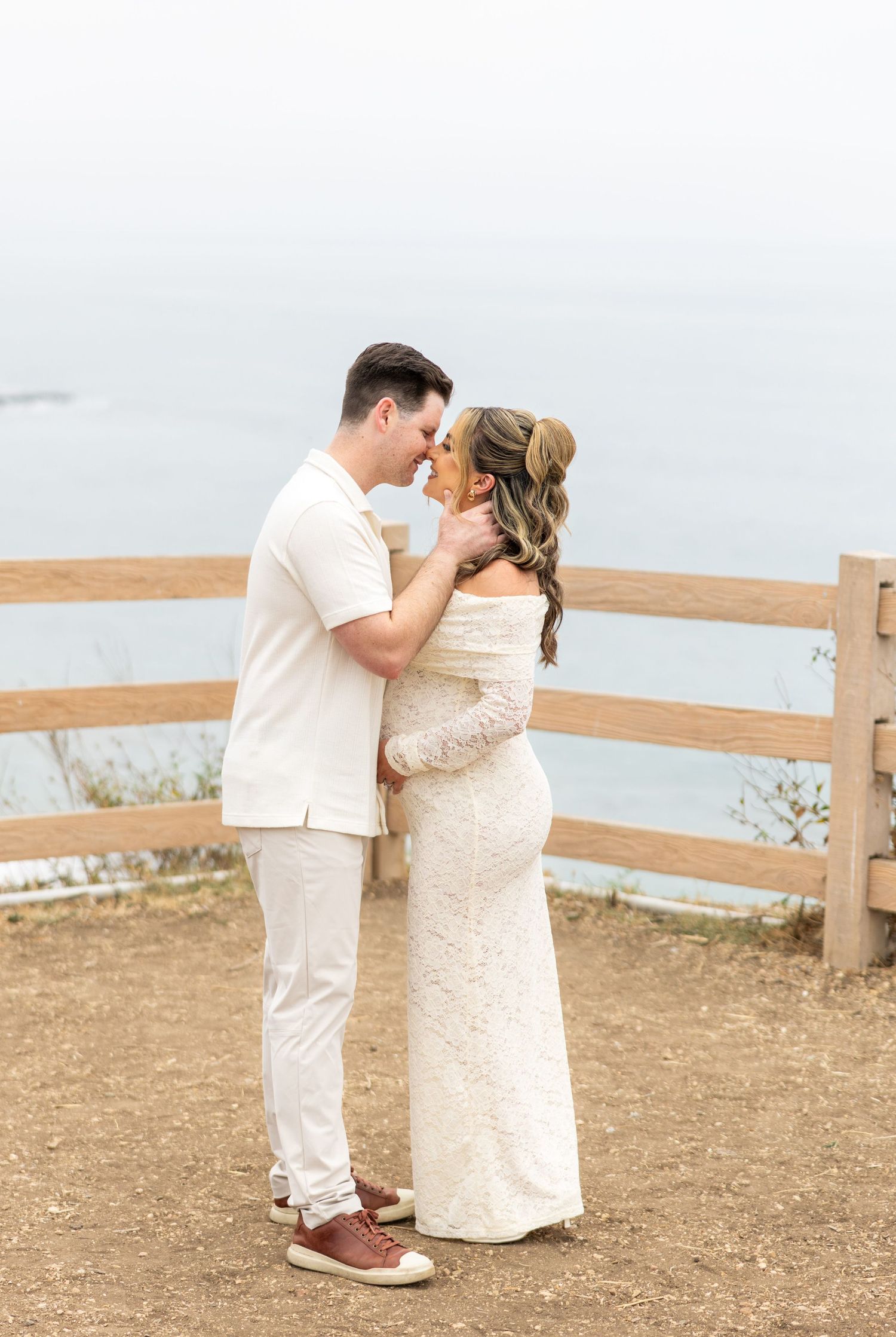 A couple shares a romantic seaside moment in matching white outfits on a hilltop path.