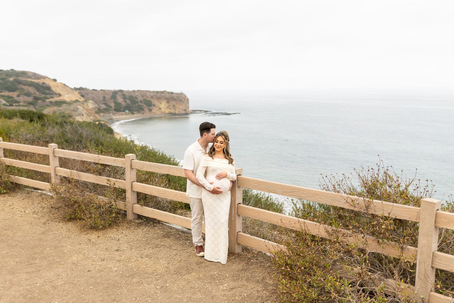 A couple shares an intimate embrace along a coastal cliffside path with wooden fence and ocean views in the background.