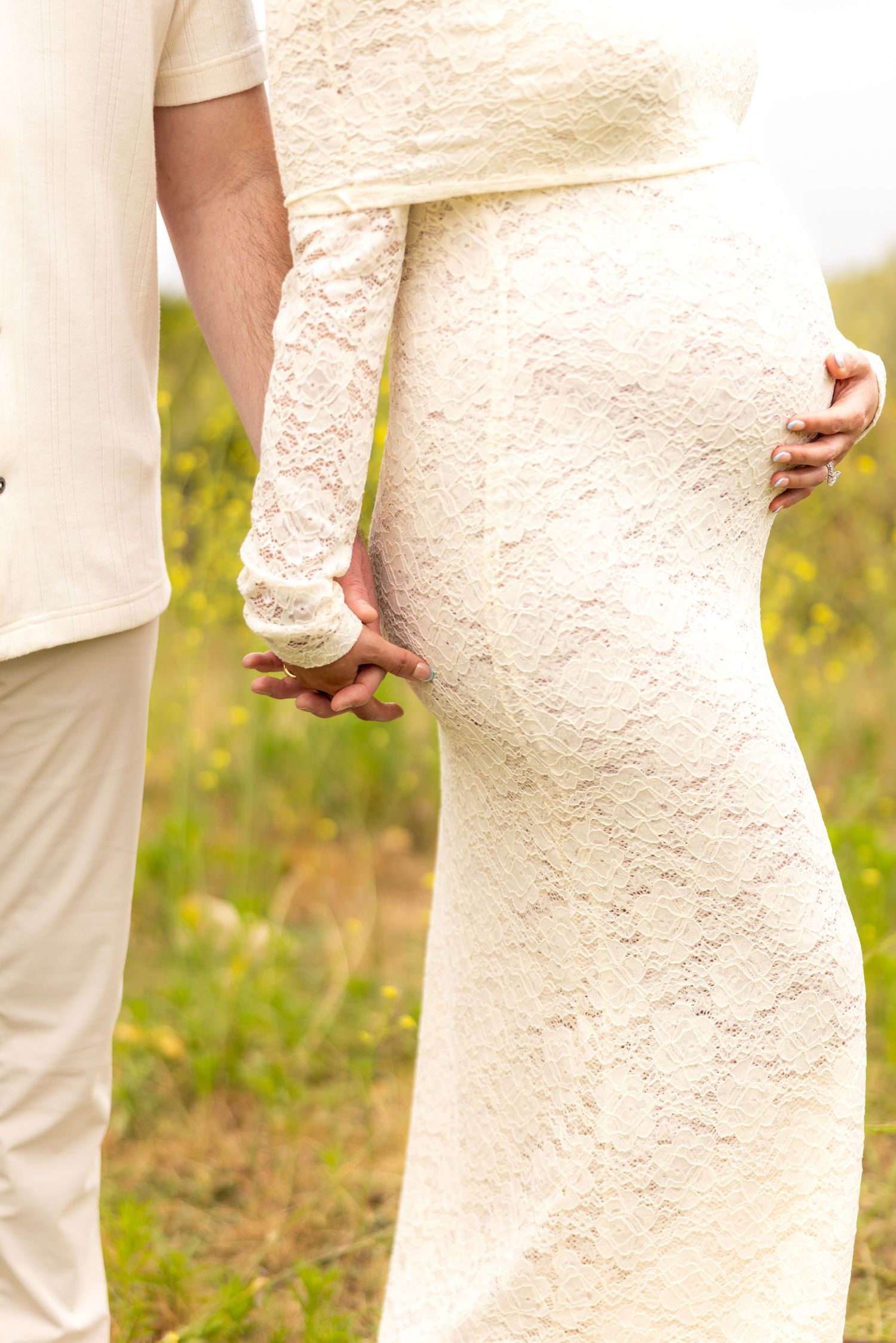 Close-up view of newlyweds holding hands outdoors with bride in lace dress and groom in light colored suit.