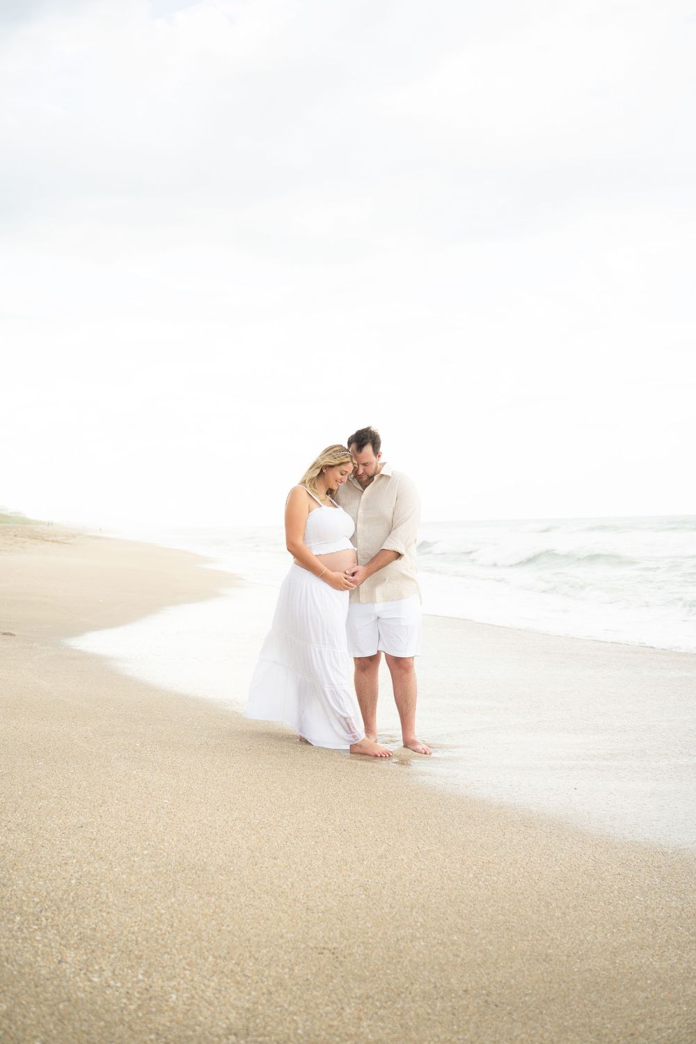 Pregnant couple in white embracing on a sandy beach shore with ocean waves in the background.