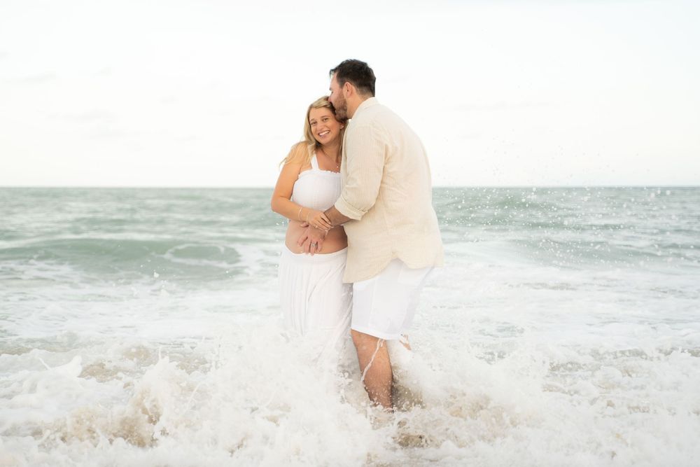 Couple embracing on beach shore with ocean waves, woman in white maternity dress, man in beige outfit, romantic photo.