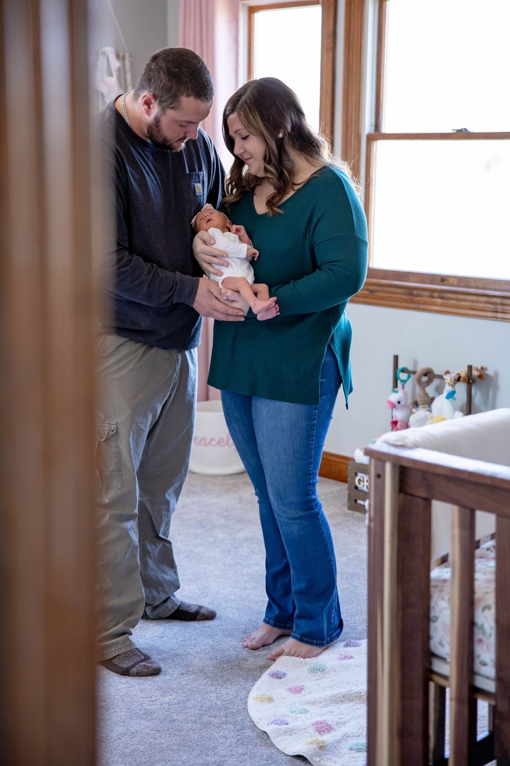 A couple stands together on a carpeted floor near windows while looking down at a baby between them. Family session. North Carolina.