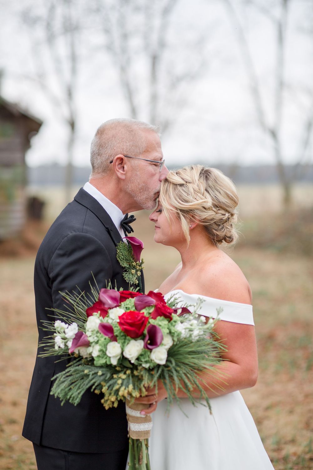 A tender wedding moment captured outdoors with a beautiful red and white bouquet featuring pine accents.