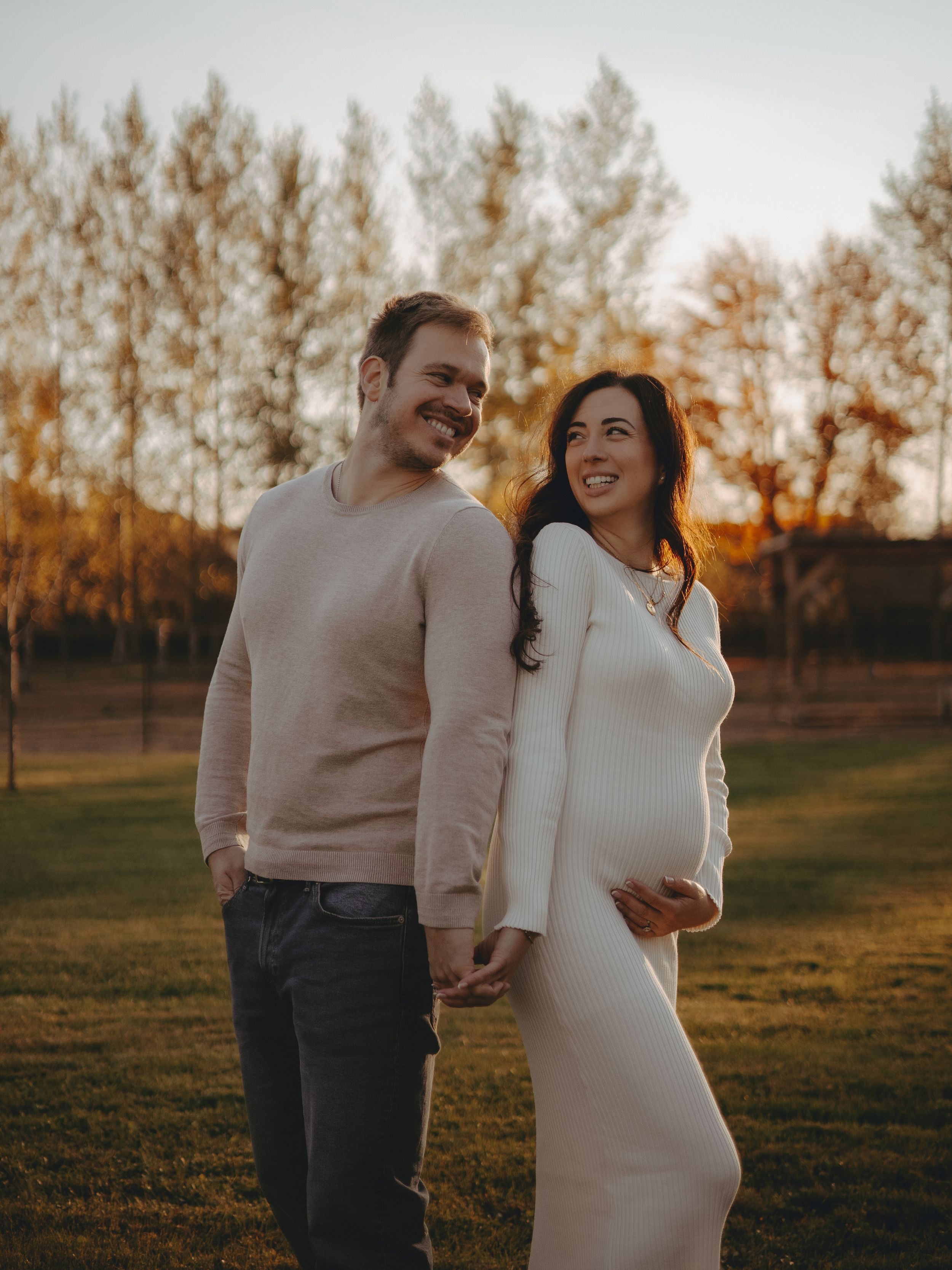 Autumn maternity photoshoot with couple in neutral colors standing in field at sunset.