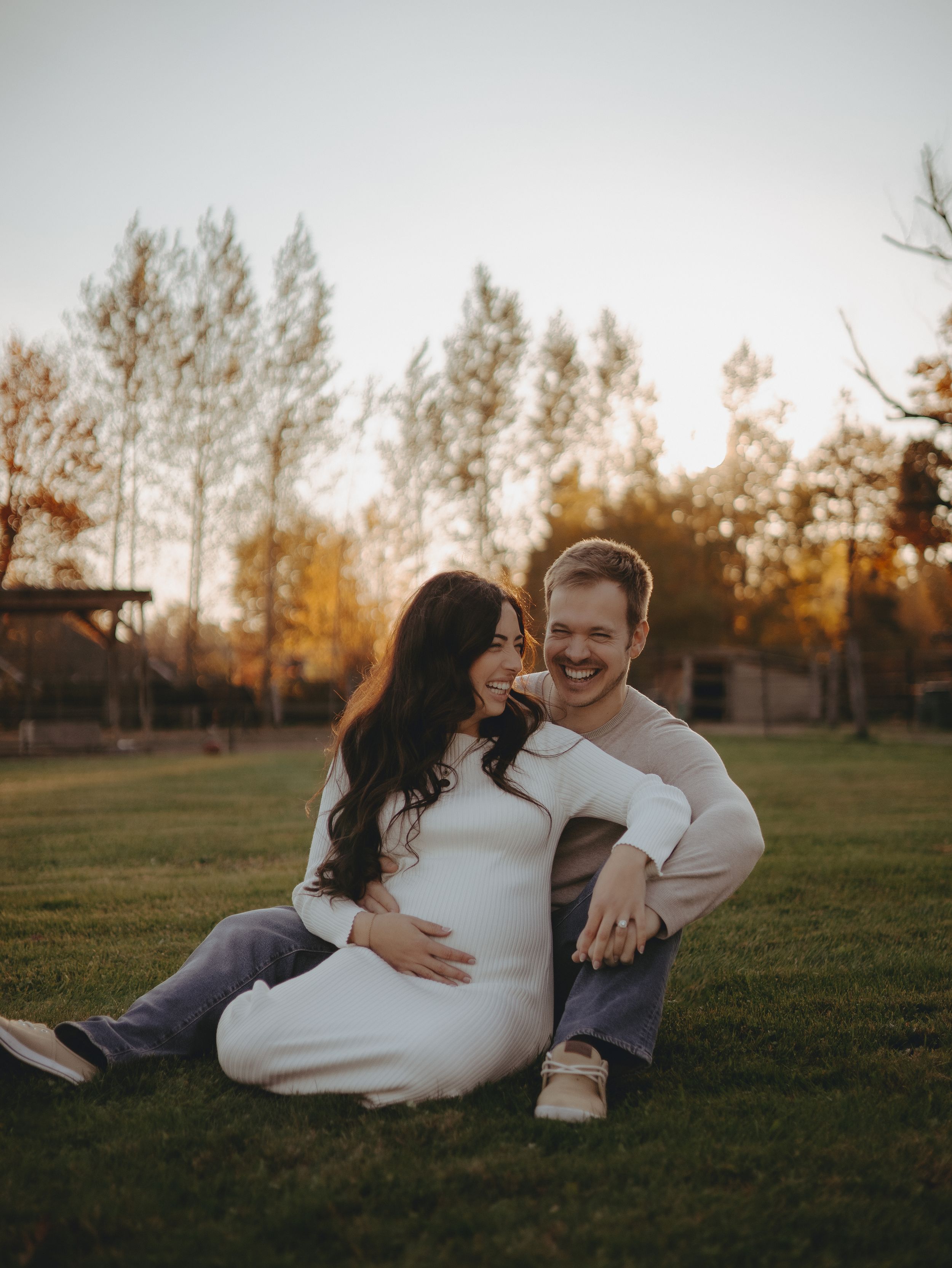 A couple sits together on grass during golden hour with trees in the background.
