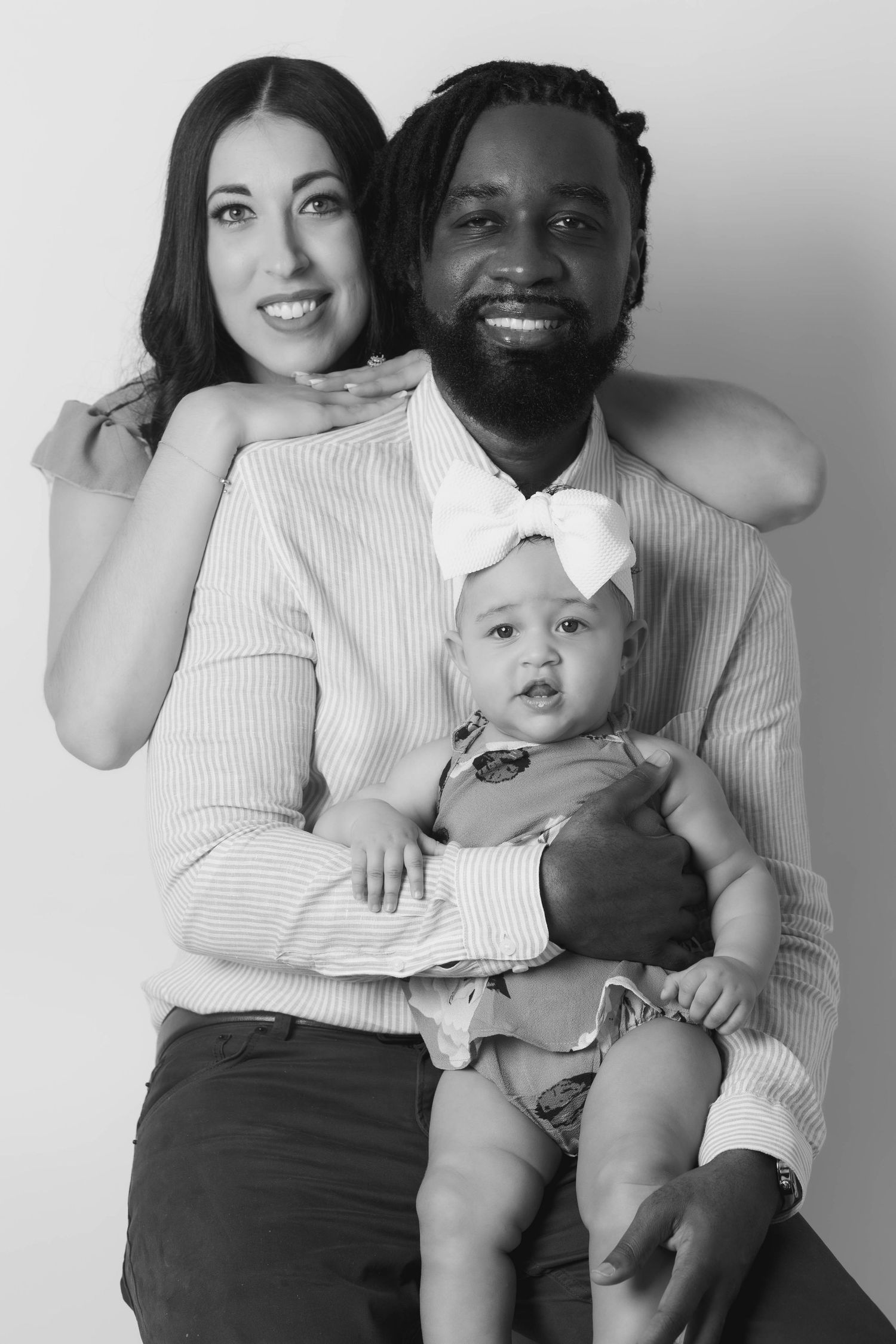A black and white family portrait featuring a couple and their baby with a bow headband.