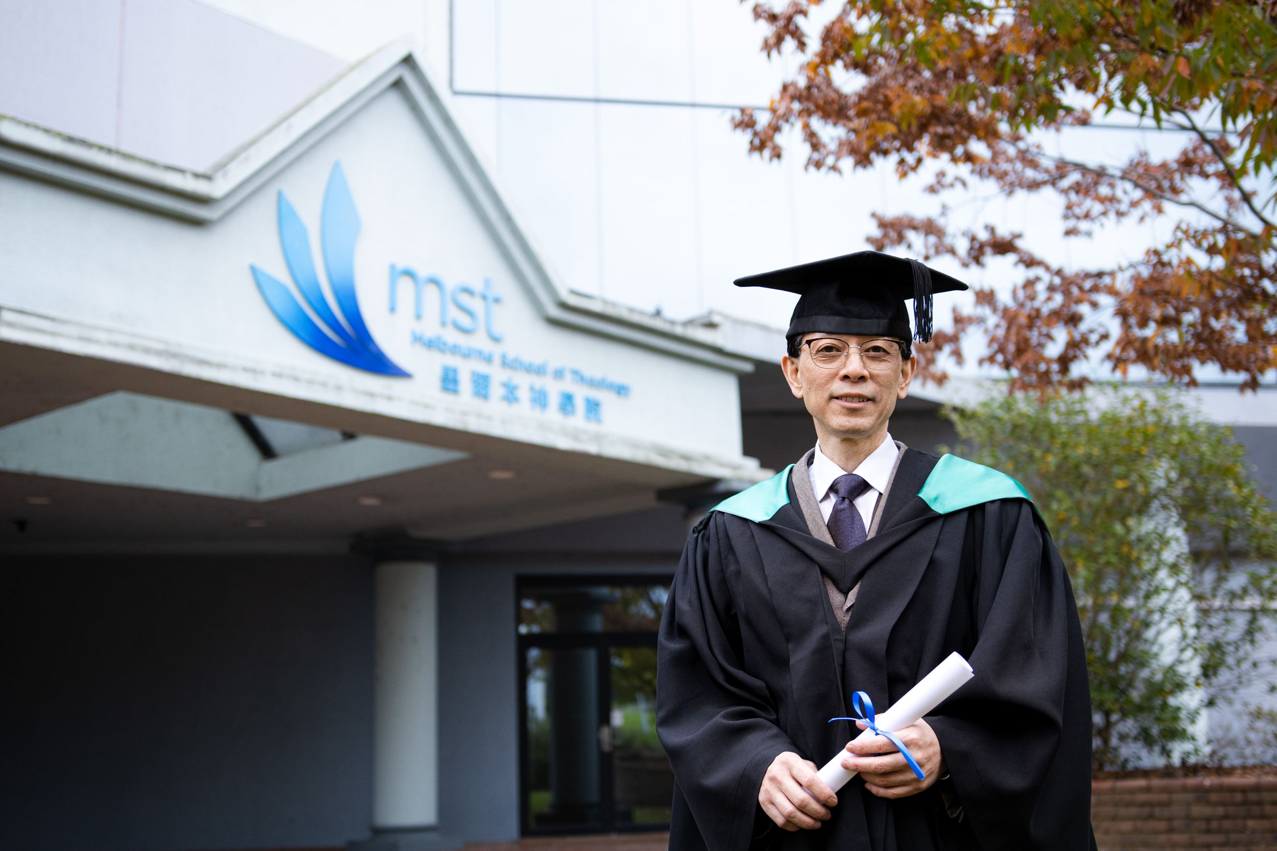 A graduate in black and green academic regalia poses with diploma outside MSE building.