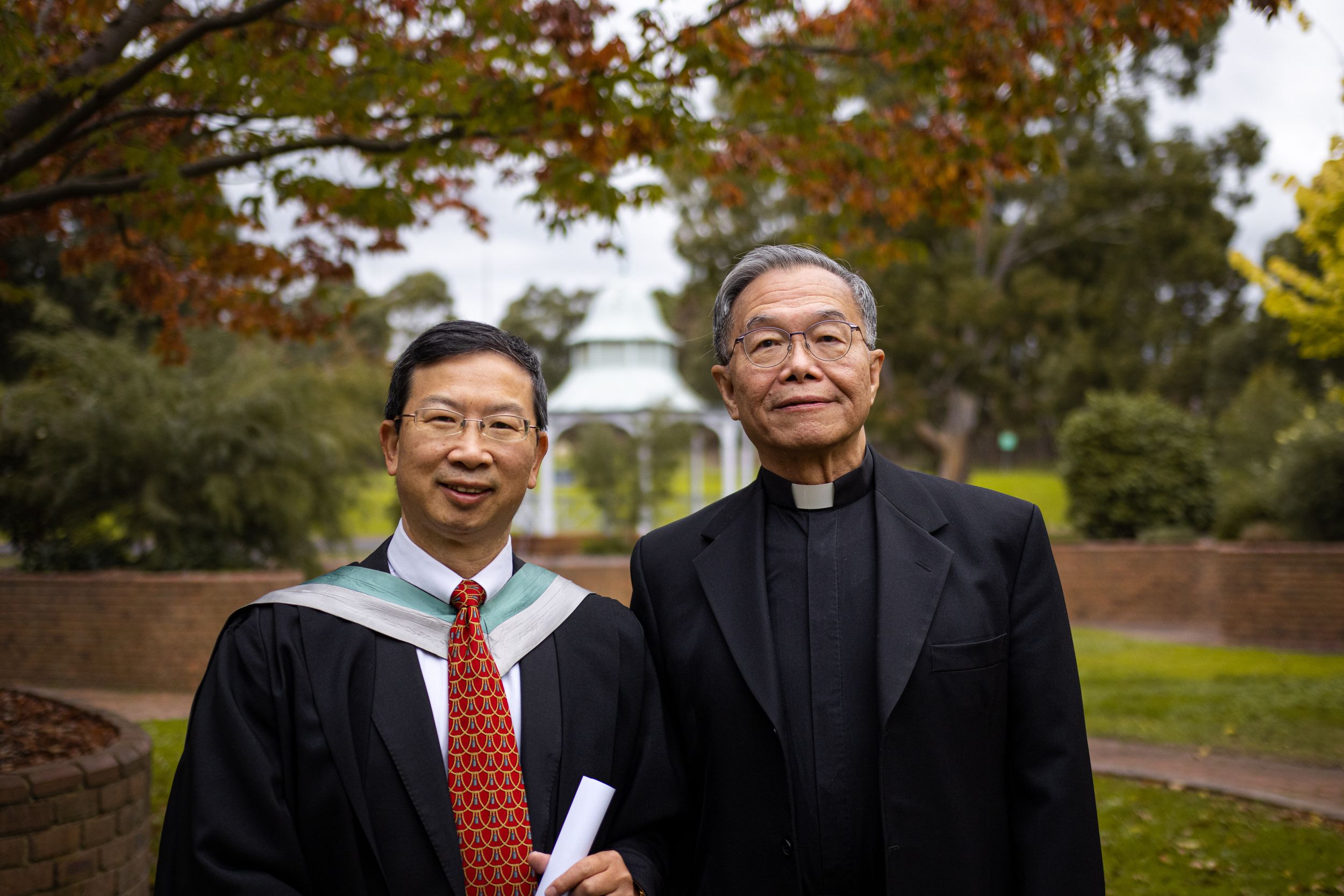Two figures in academic and religious attire stand together on campus grounds.