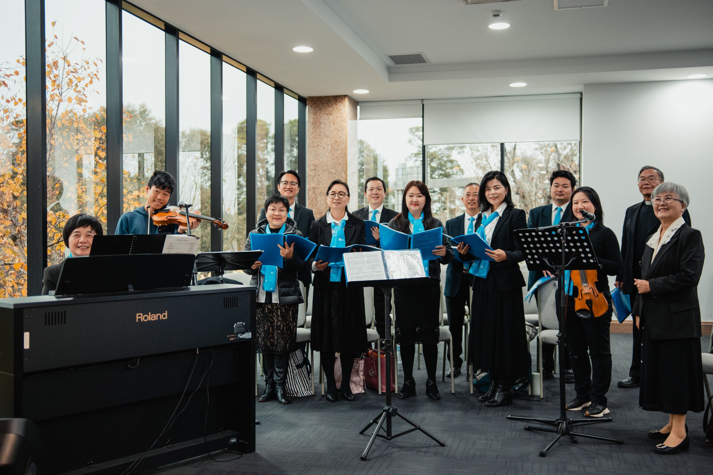A choir group performing in a bright room with large windows and a piano.