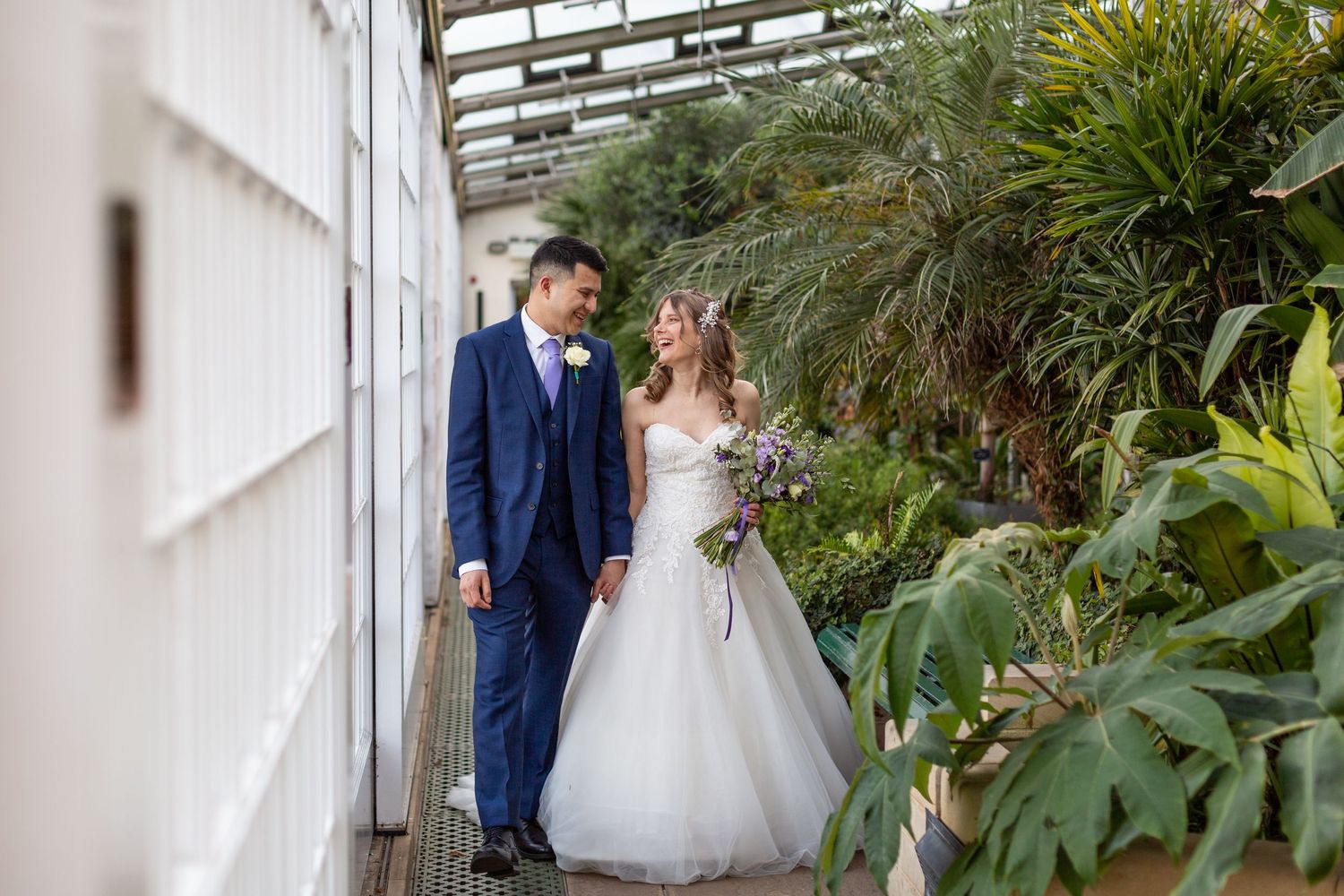 Couple in wedding attire walks through lush greenhouse corridor.