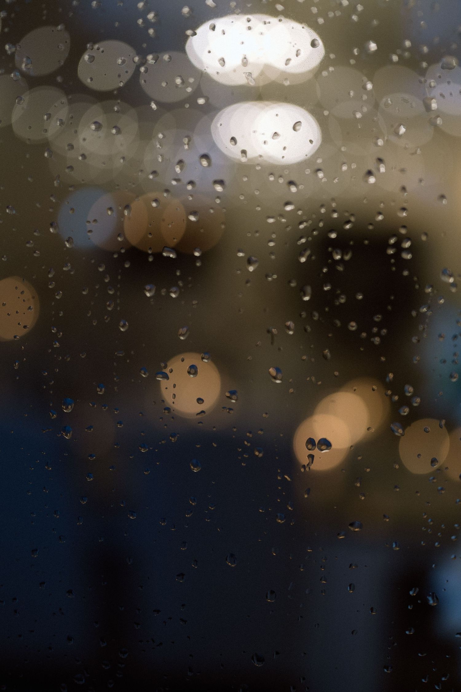 Raindrops on a window pane create bokeh effects with blurred lights in the background.