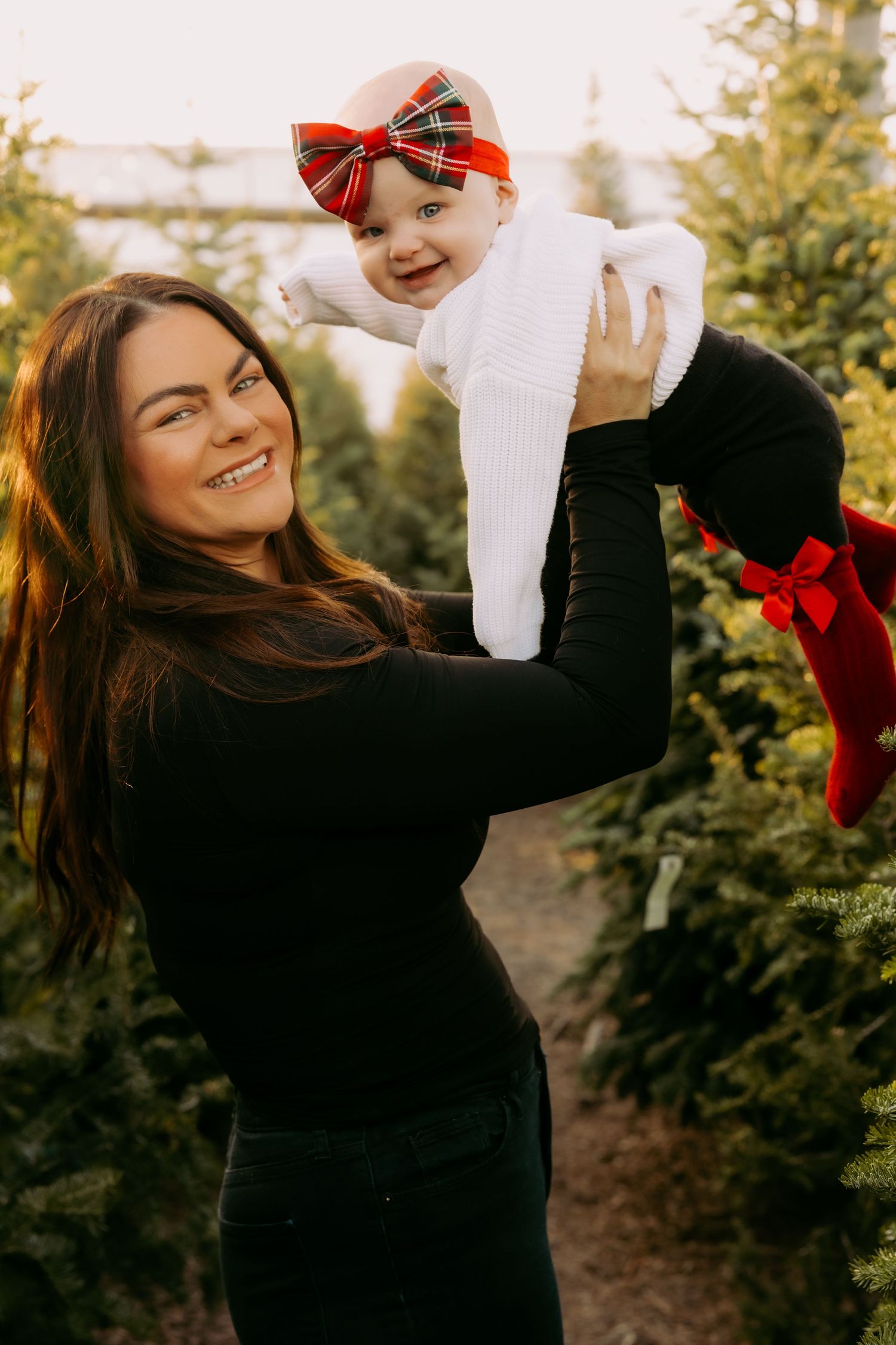 A joyful outdoor autumn portrait featuring a black-clad woman lifting a baby wearing a red bow and white sweater.