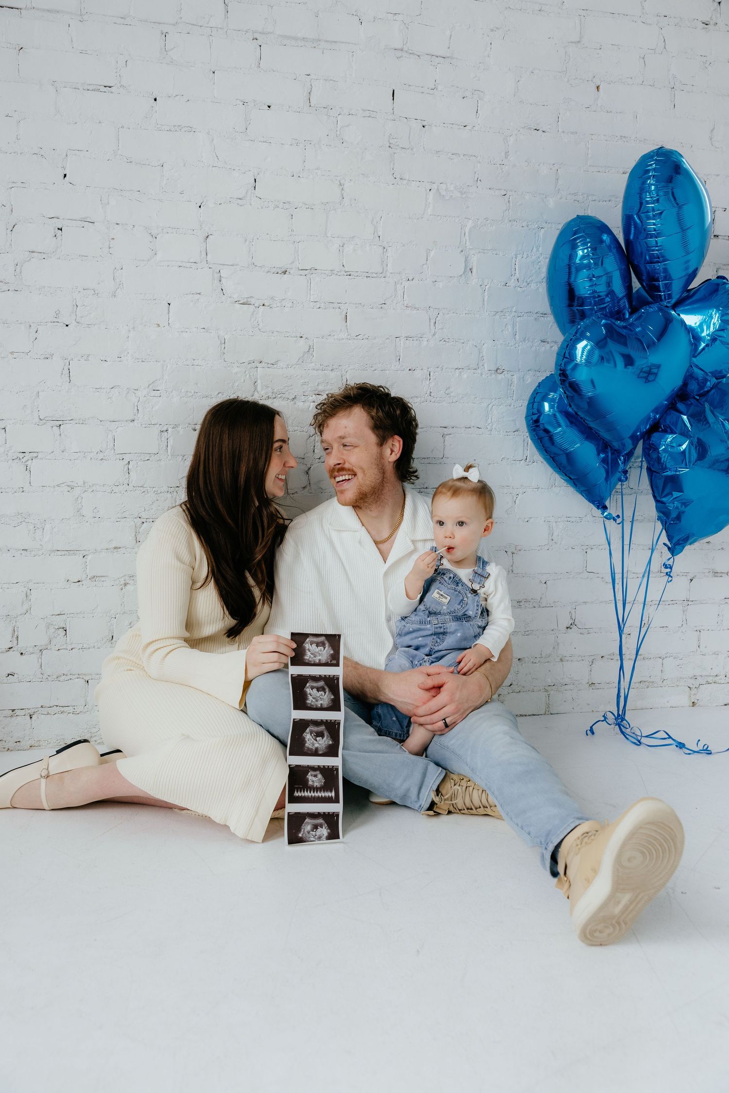 Family sits against white brick wall with blue balloons and ultrasound photos sharing a joyful pregnancy announcement moment.
