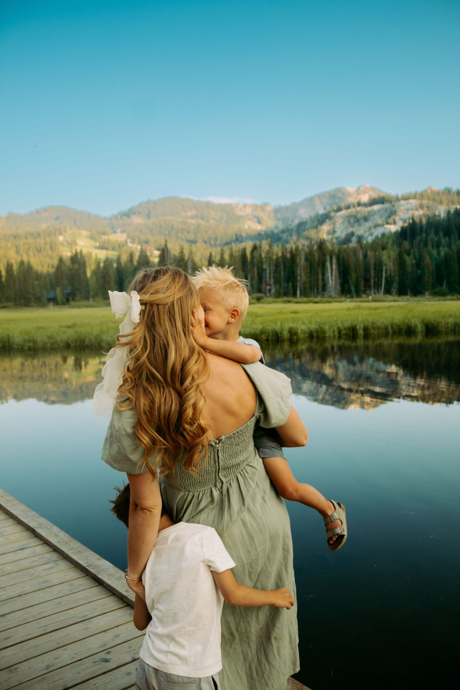 Mother holding child on dock with mountain lake and forest backdrop at sunset.