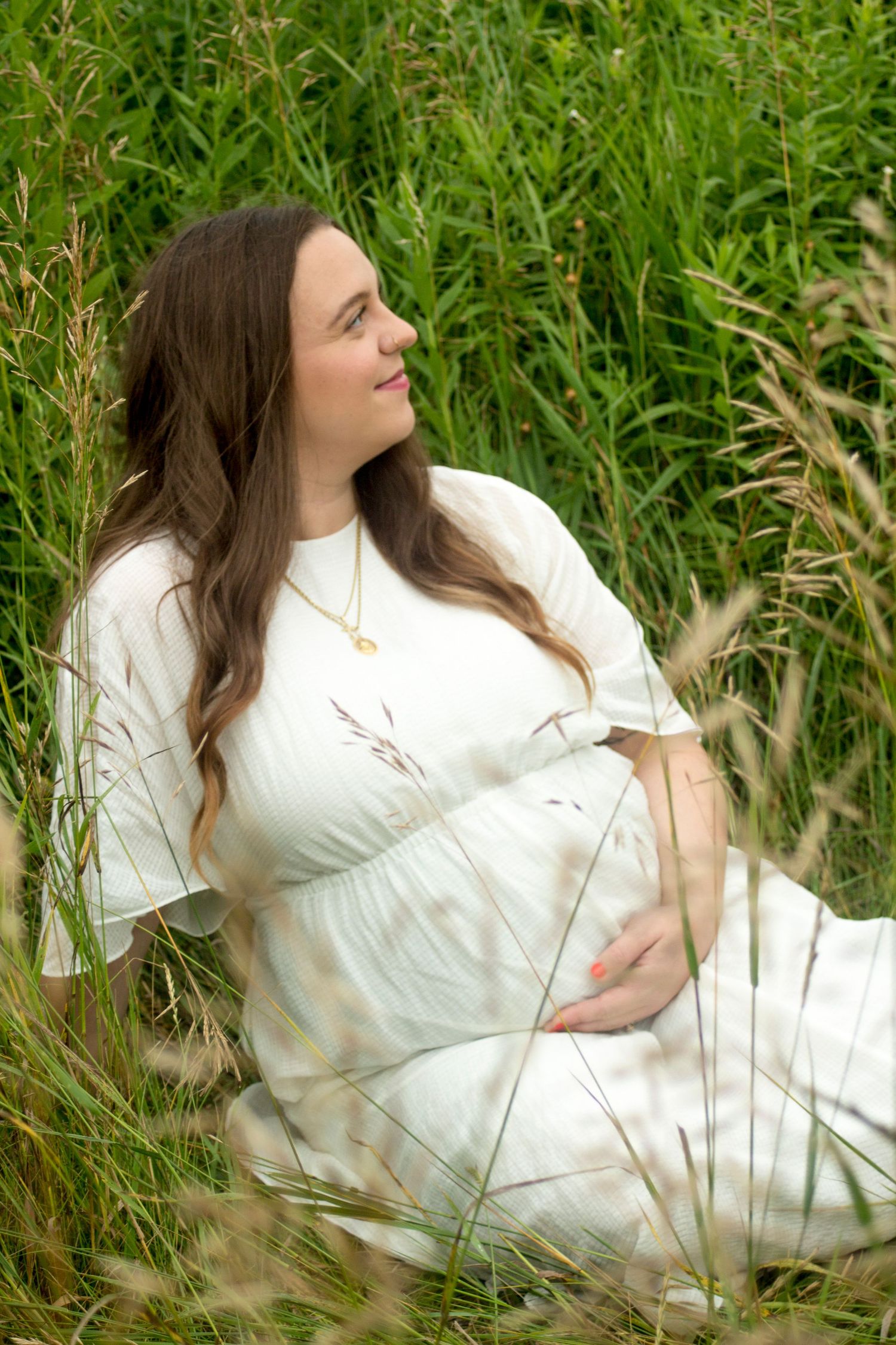 Maternity photo of a women relaxed among tall grass holding her baby bump and looking up.