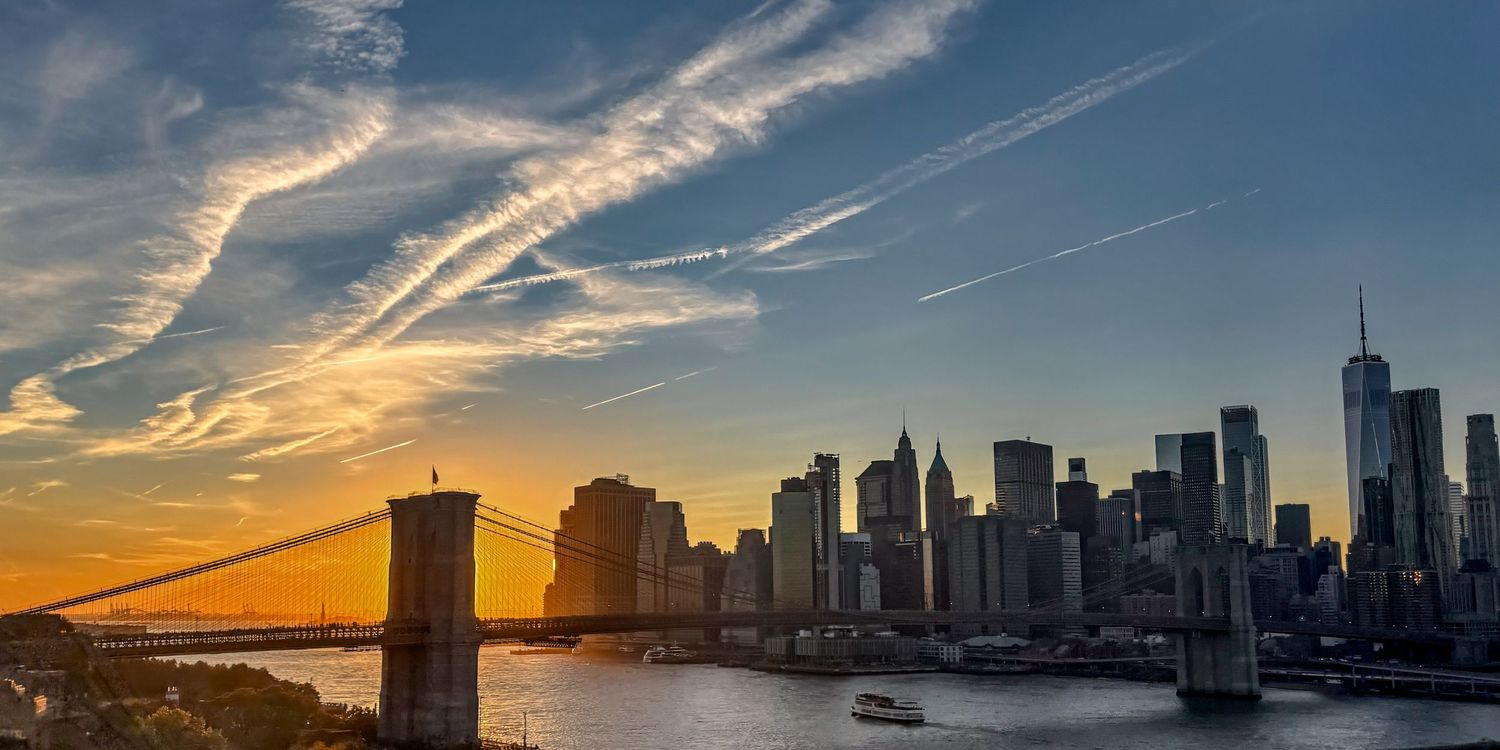 The Brooklyn Bridge and Manhattan skyline silhouetted against a dramatic sunset sky with streaking clouds.