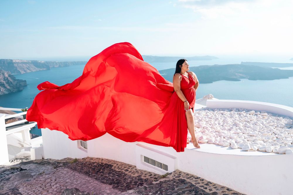 Billowing red dress catches wind on white terrace overlooking Mediterranean Sea.