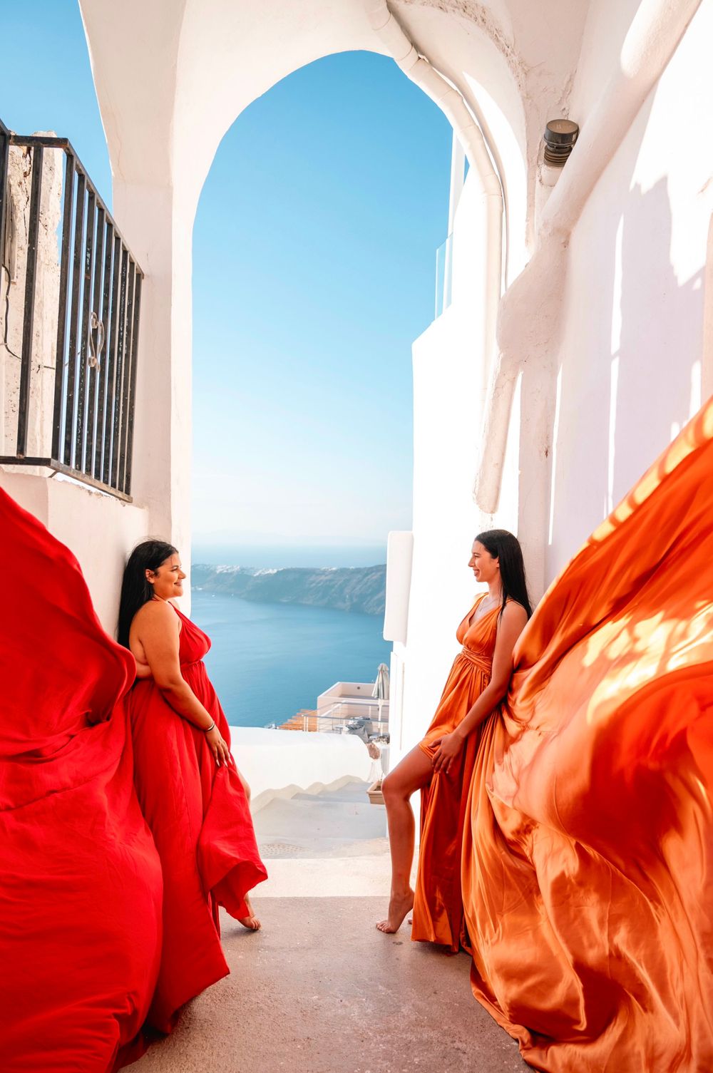 Two flowing dresses in red and orange create dramatic movement against white Mediterranean architecture.