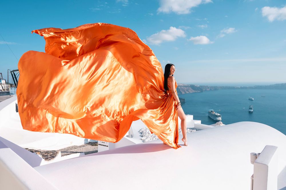Orange dress fabric flows in wind on white Santorini rooftop with ocean backdrop.