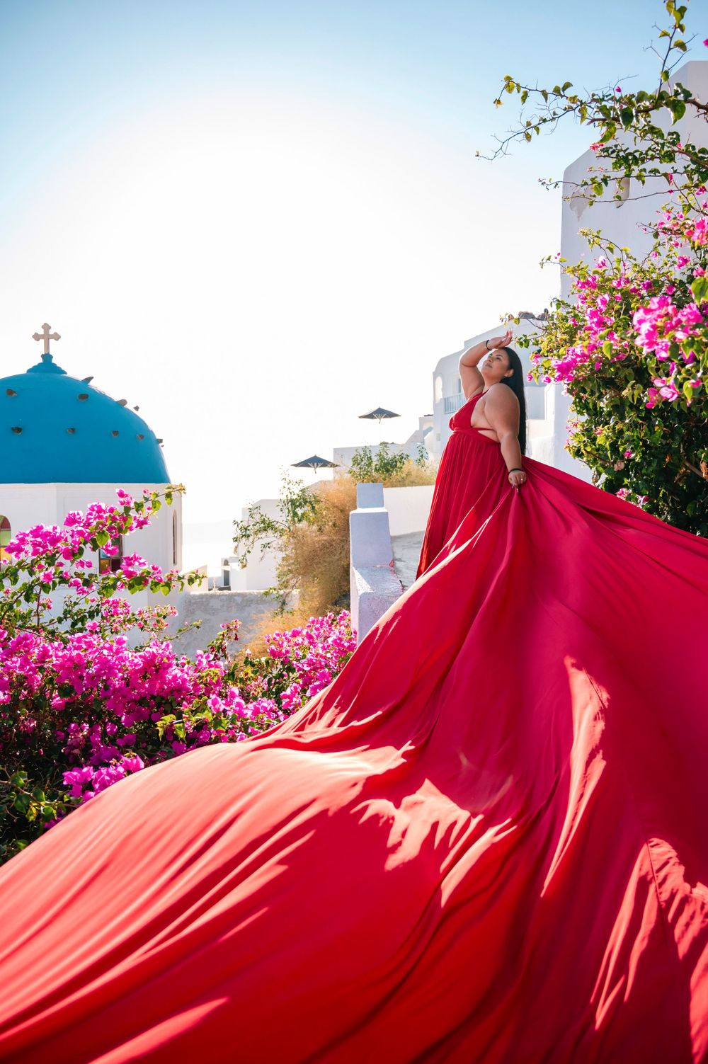 Flowing red dress billows dramatically against white Greek architecture and pink bougainvillea in Santorini.