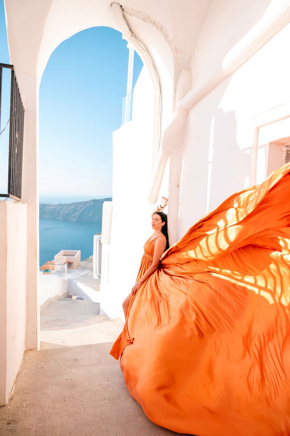 Billowing orange dress creates dramatic movement against white Mediterranean archways and blue sea views.
