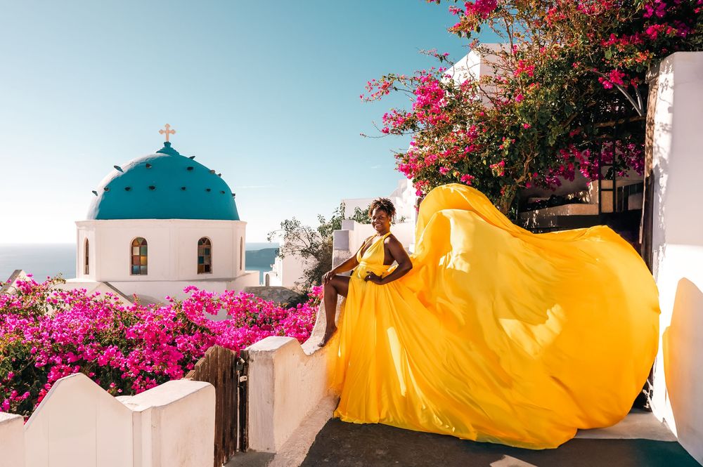 Woman in flowing yellow gown poses in Santorini Greece with blue domed church and pink bougainvillea.