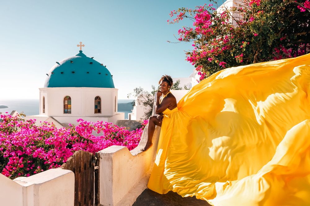 Woman in flowing yellow dress poses on wall in Santorini with blue-domed church and pink bougainvillea.